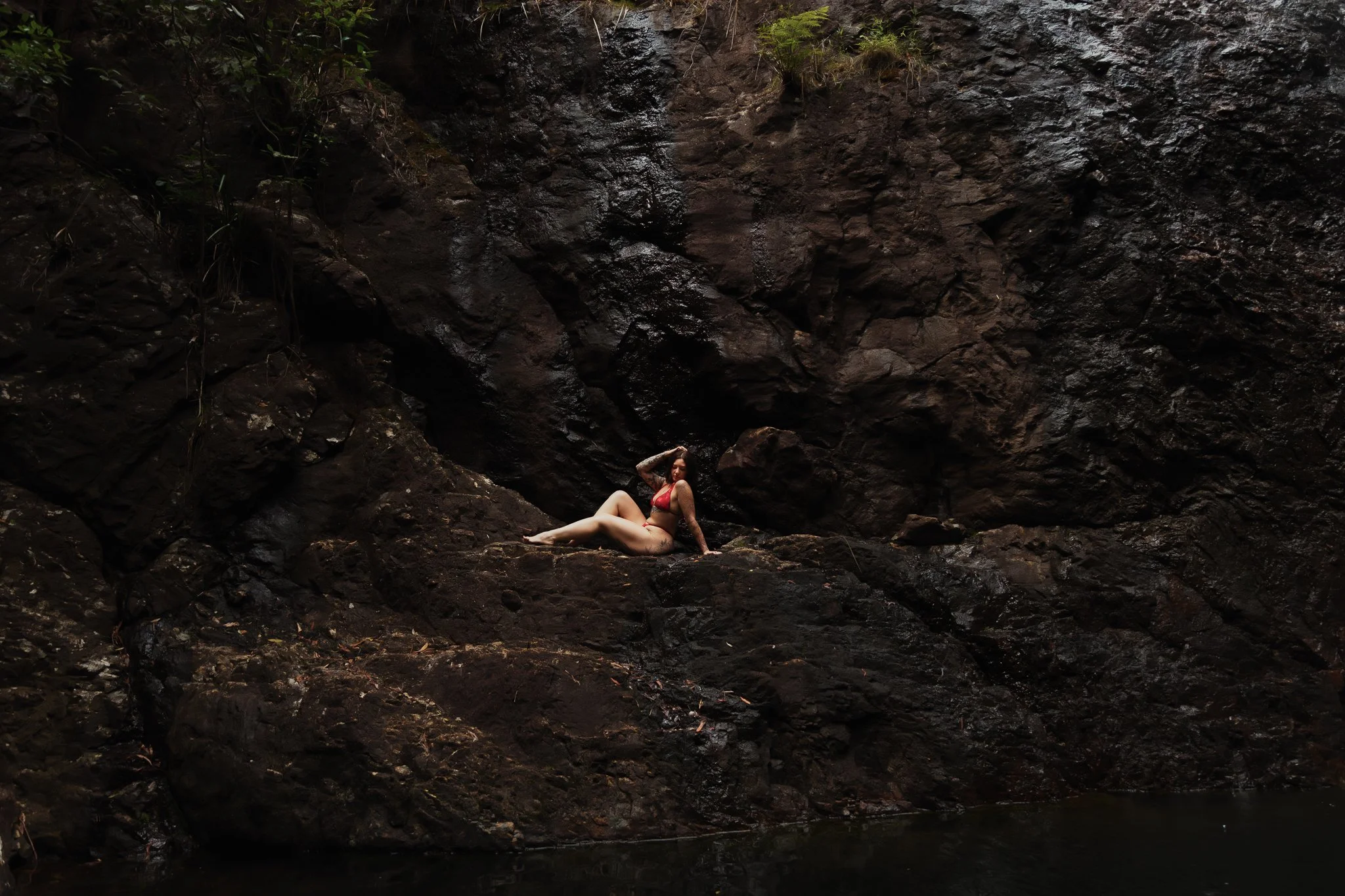 A woman in a red bikini lying on a large rock beside a body of water, surrounded by dark, wet rocks and foliage.