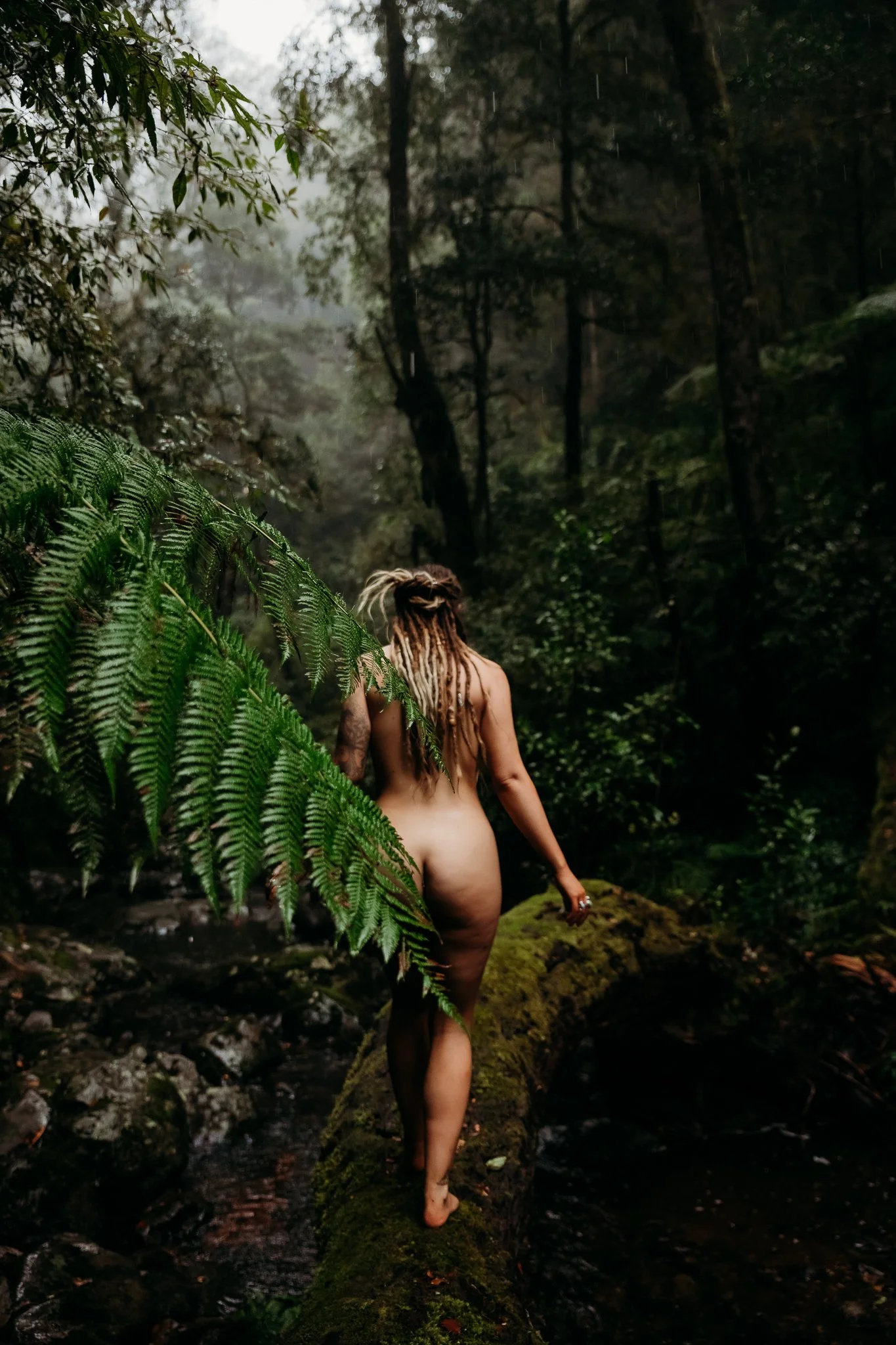 A nude woman with dreadlocks walking on a moss-covered log through a dense, foggy forest.
