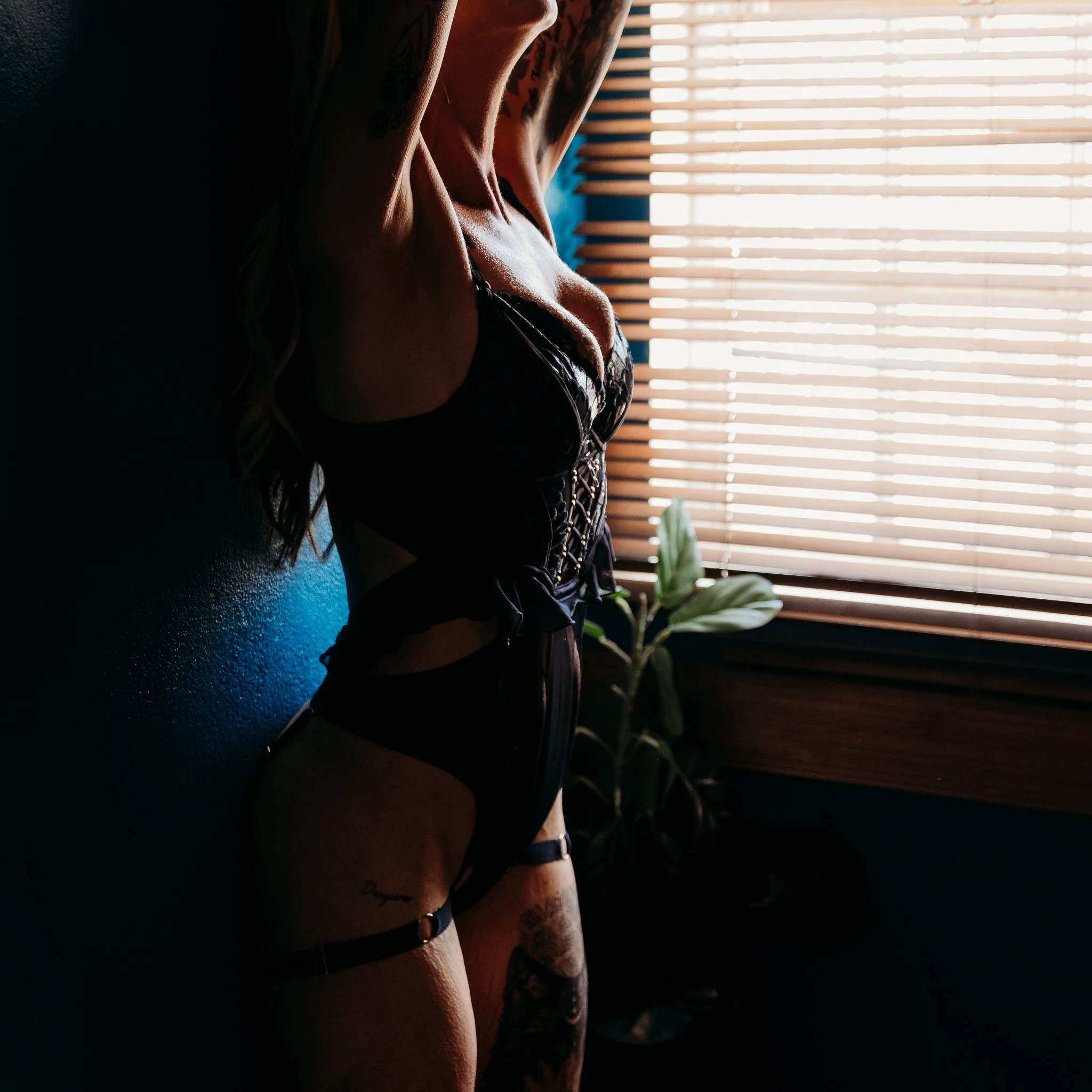 A person with tattoos and a black wristwatch sitting near a window with closed blinds, with natural light streaming through.
