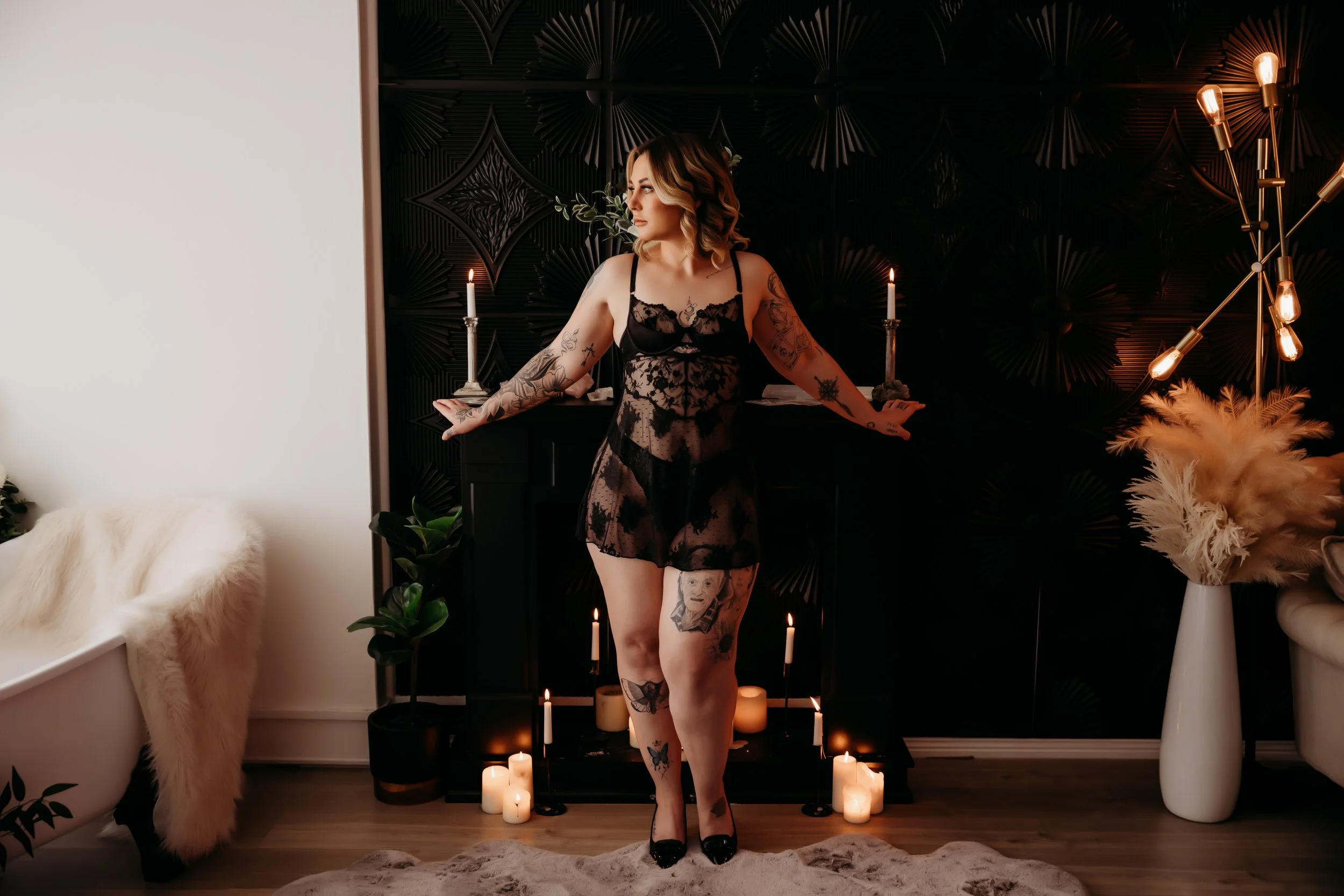 A woman with tattoos wearing black lace lingerie and high heels standing in front of a fireplace with candles and decorative plants in a dimly lit room.