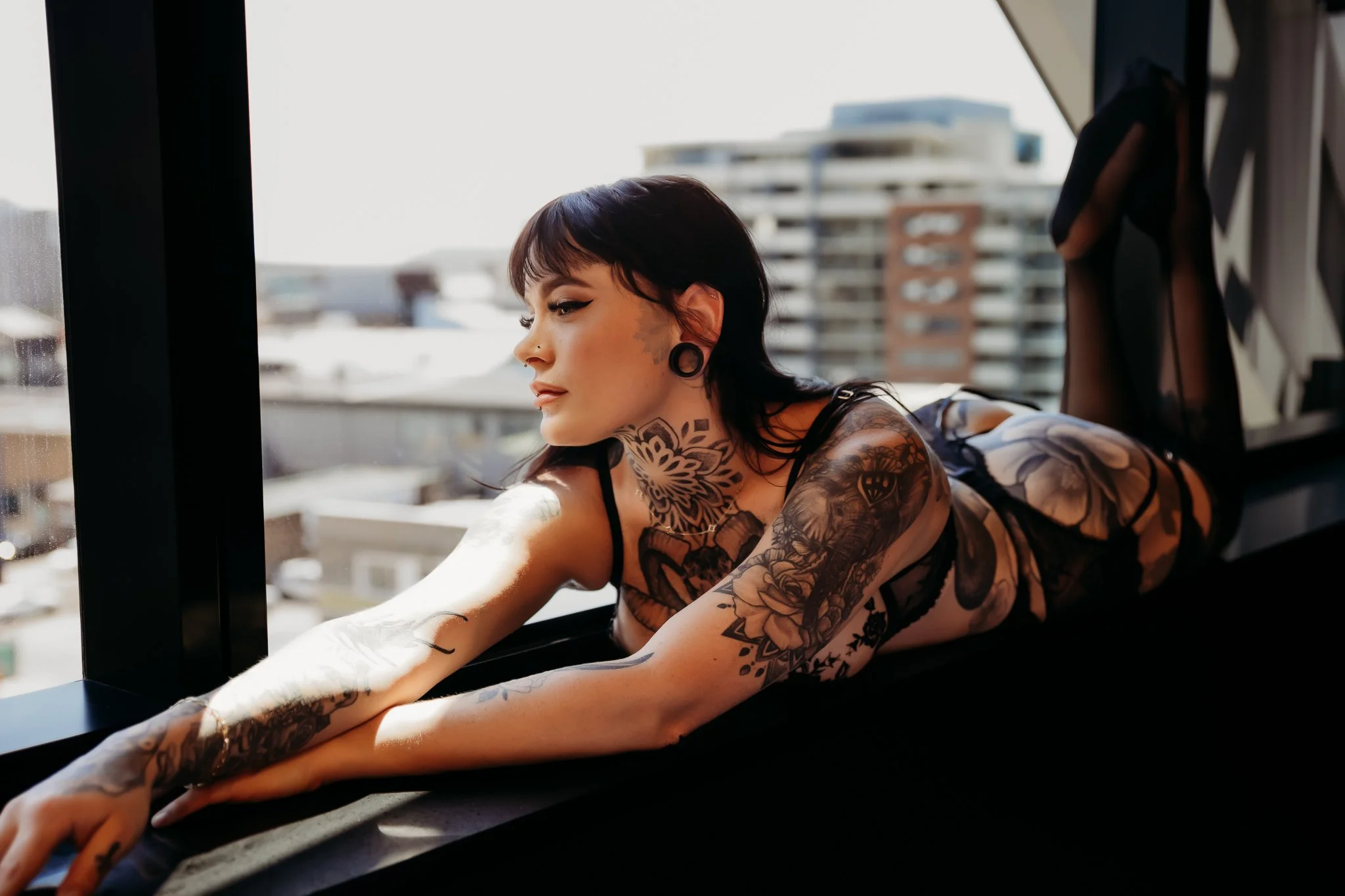 Tattooed woman lying on her stomach on a windowsill in an urban apartment, gazing out the window with city buildings in the background.