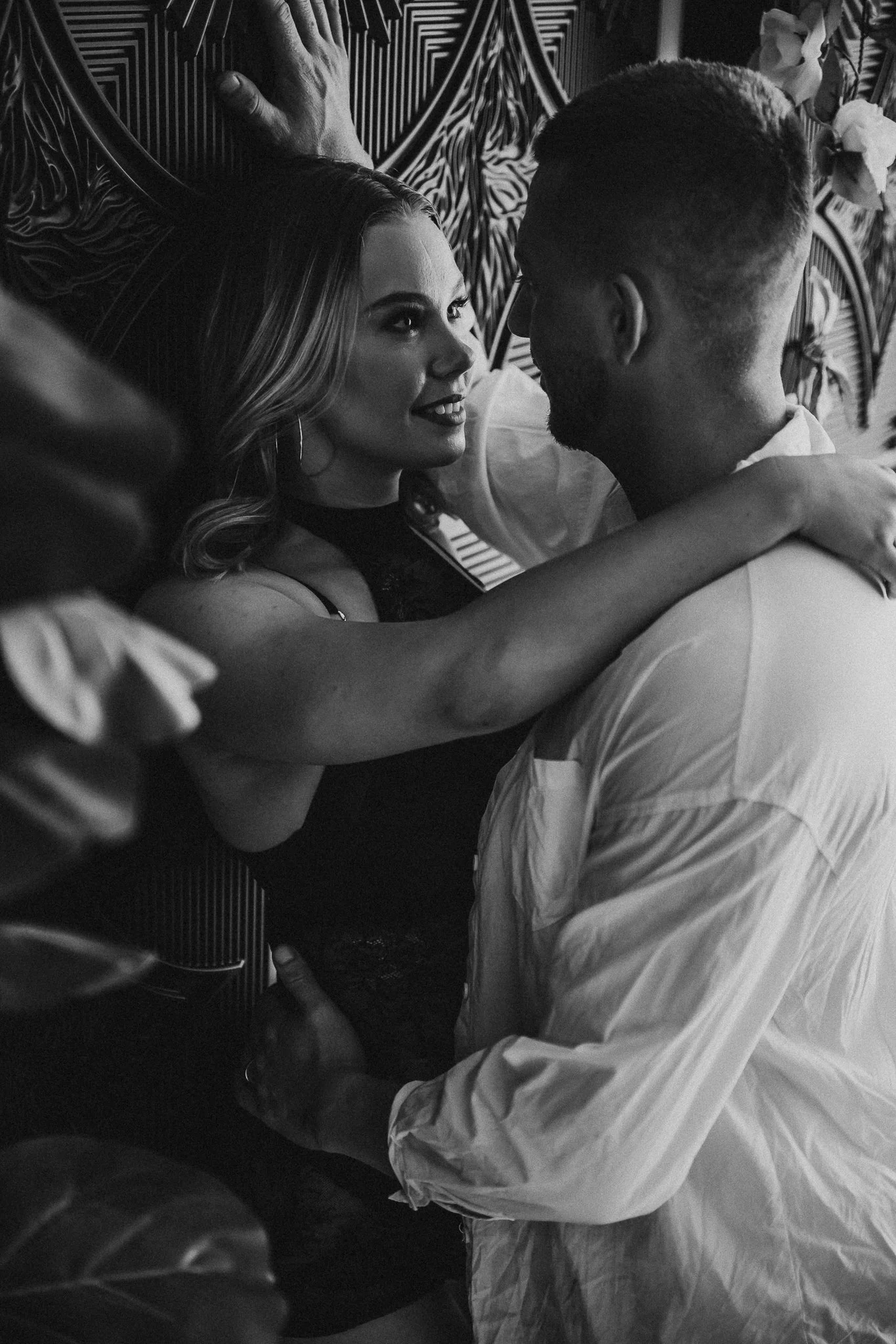 A black-and-white photo of a woman and man close-up, looking into each other's eyes, with the woman smiling and the man gently holding her, and an arm partially visible in the foreground.