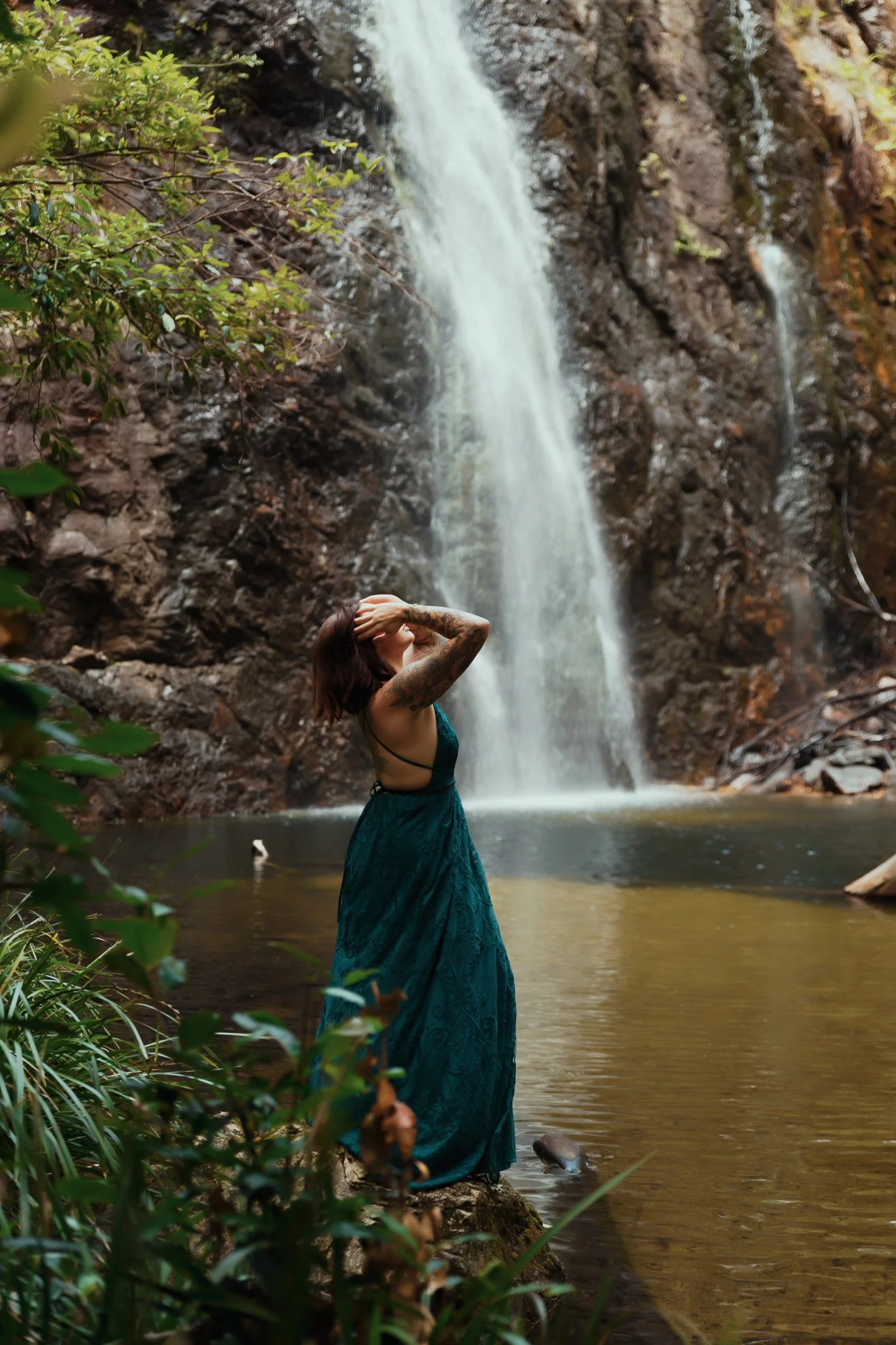 A woman in a teal dress stands near a waterfall in a forested area, with her hands on her head.