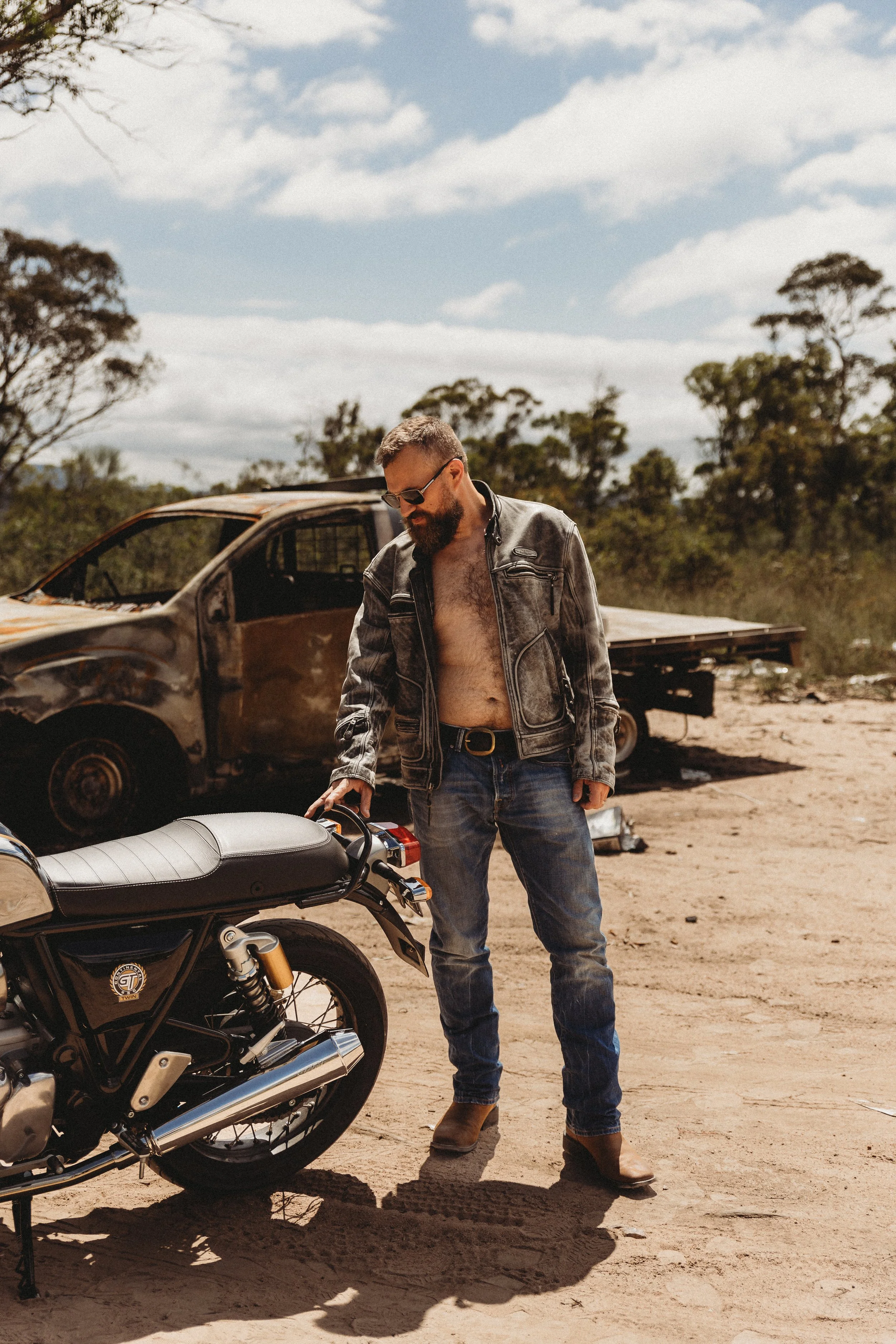 A man with a beard, glasses, and a partially unbuttoned leather jacket, standing outdoors on dirt ground with a motorcycle, a rusty car, and trees in the background under a partly cloudy sky.