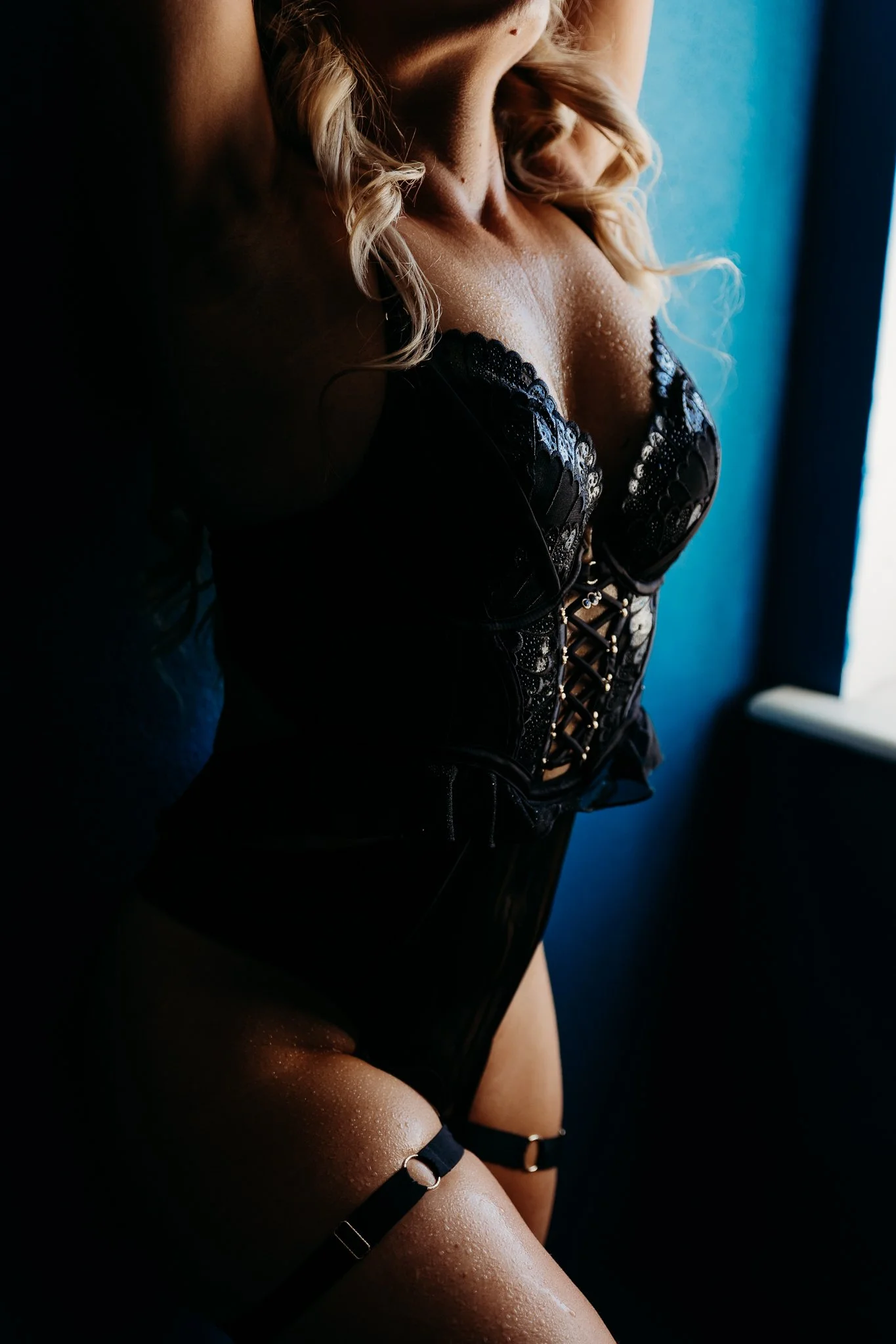A woman with lightly curled blonde hair wearing black lace lingerie, standing near a window with light illuminating her, and water droplets on her skin.