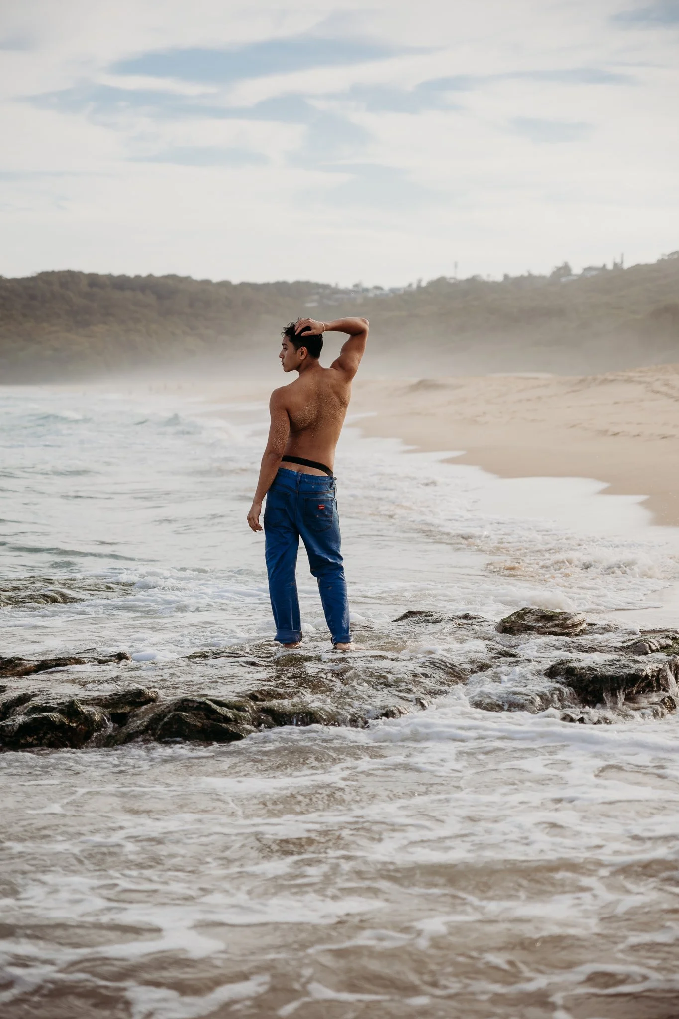 A shirtless man in blue jeans standing in the ocean waves at the beach, facing left with one hand on his head.