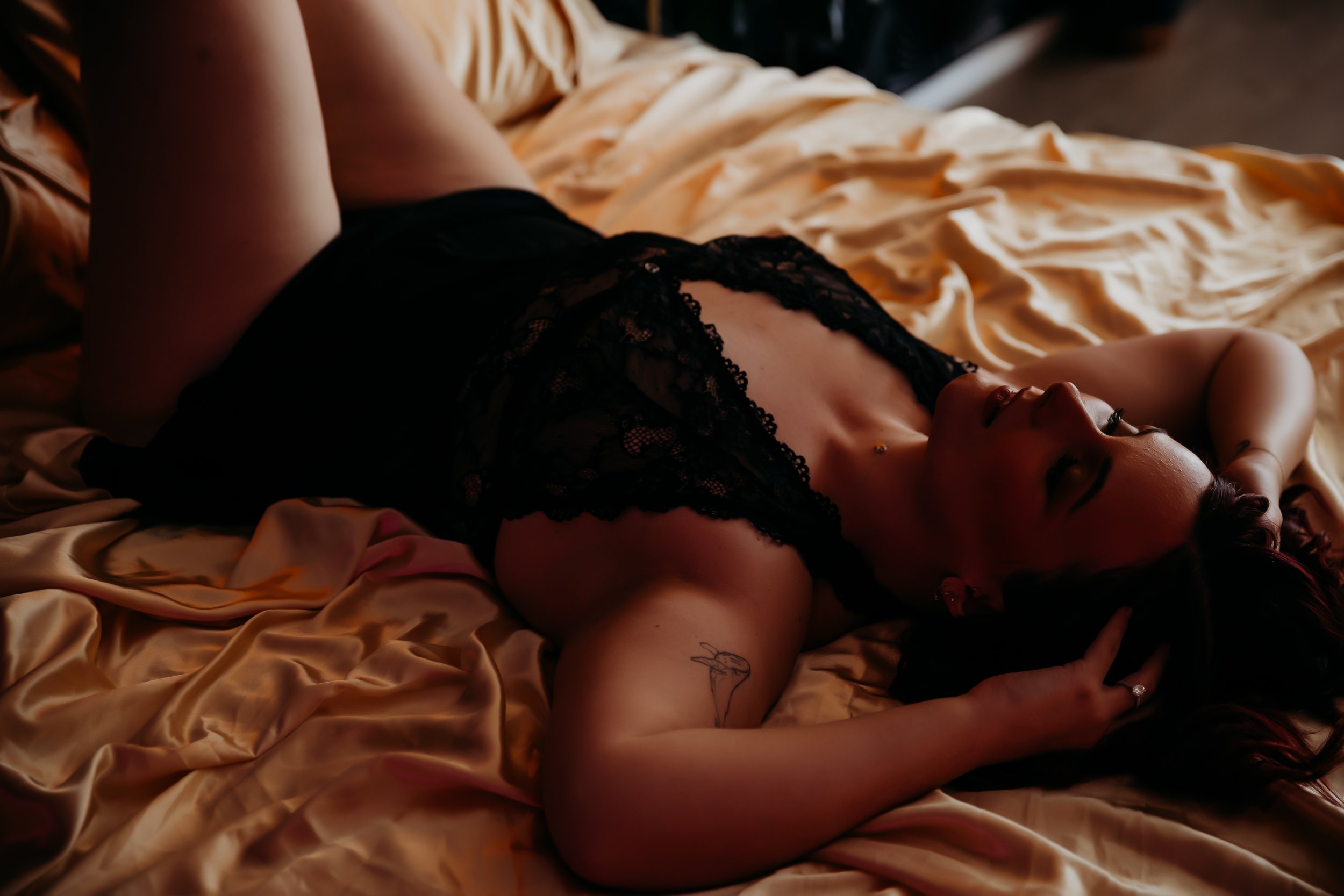 A woman lying on a bed with golden satin sheets, wearing a black lace dress, with her hair down and her eyes closed.
