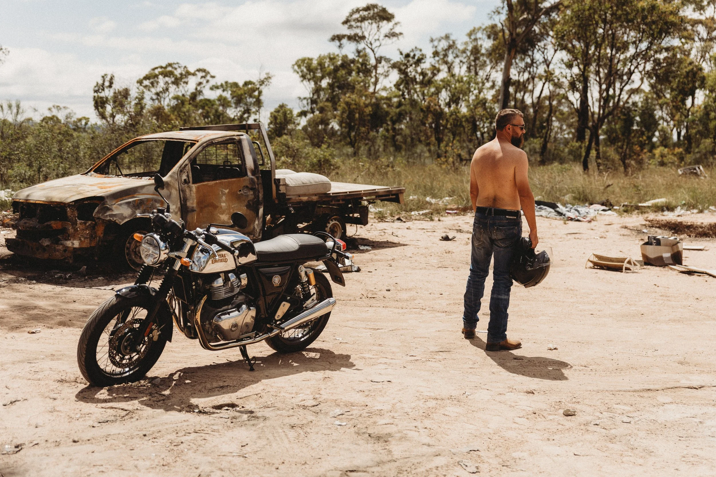 A shirtless man holding a motorcycle helmet standing next to a black motorcycle in a barren, sandy area with an old, burnt-out truck in the background and scattered debris.