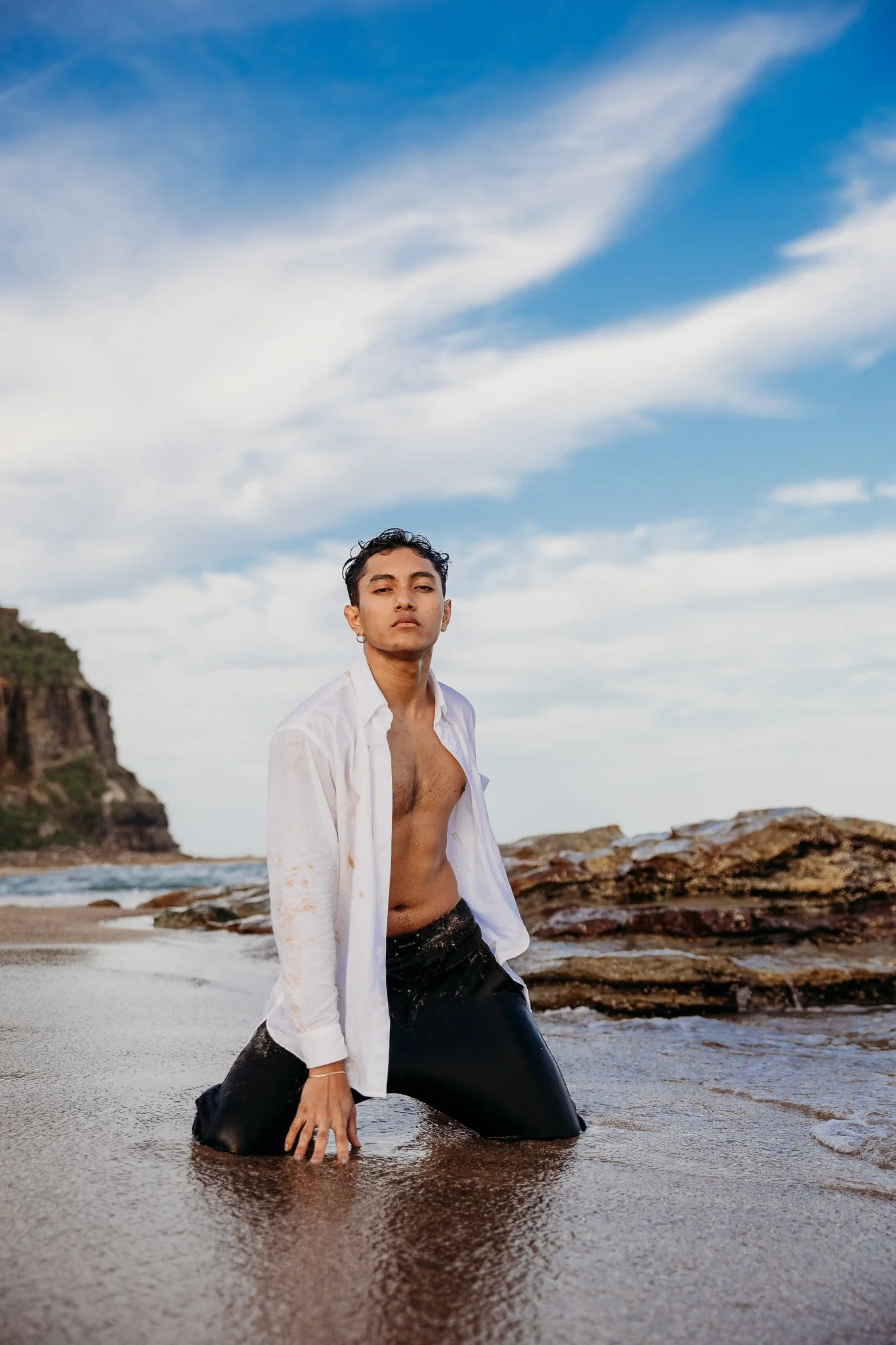 A young man kneels in shallow water on a beach, wearing a white open shirt and black pants, with a dramatic sky in the background.