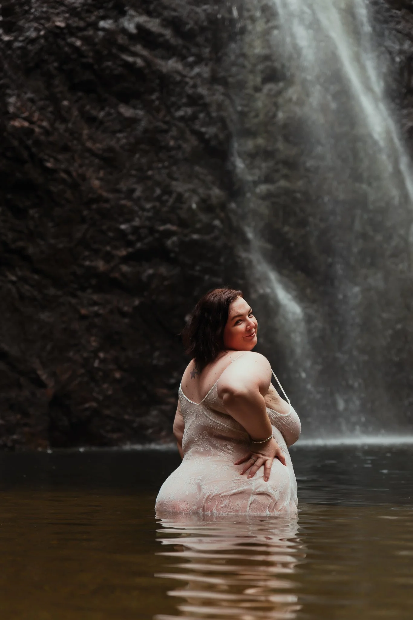 A woman standing in a waterfall with water up to her waist, with a rocky cliff and waterfall in the background.