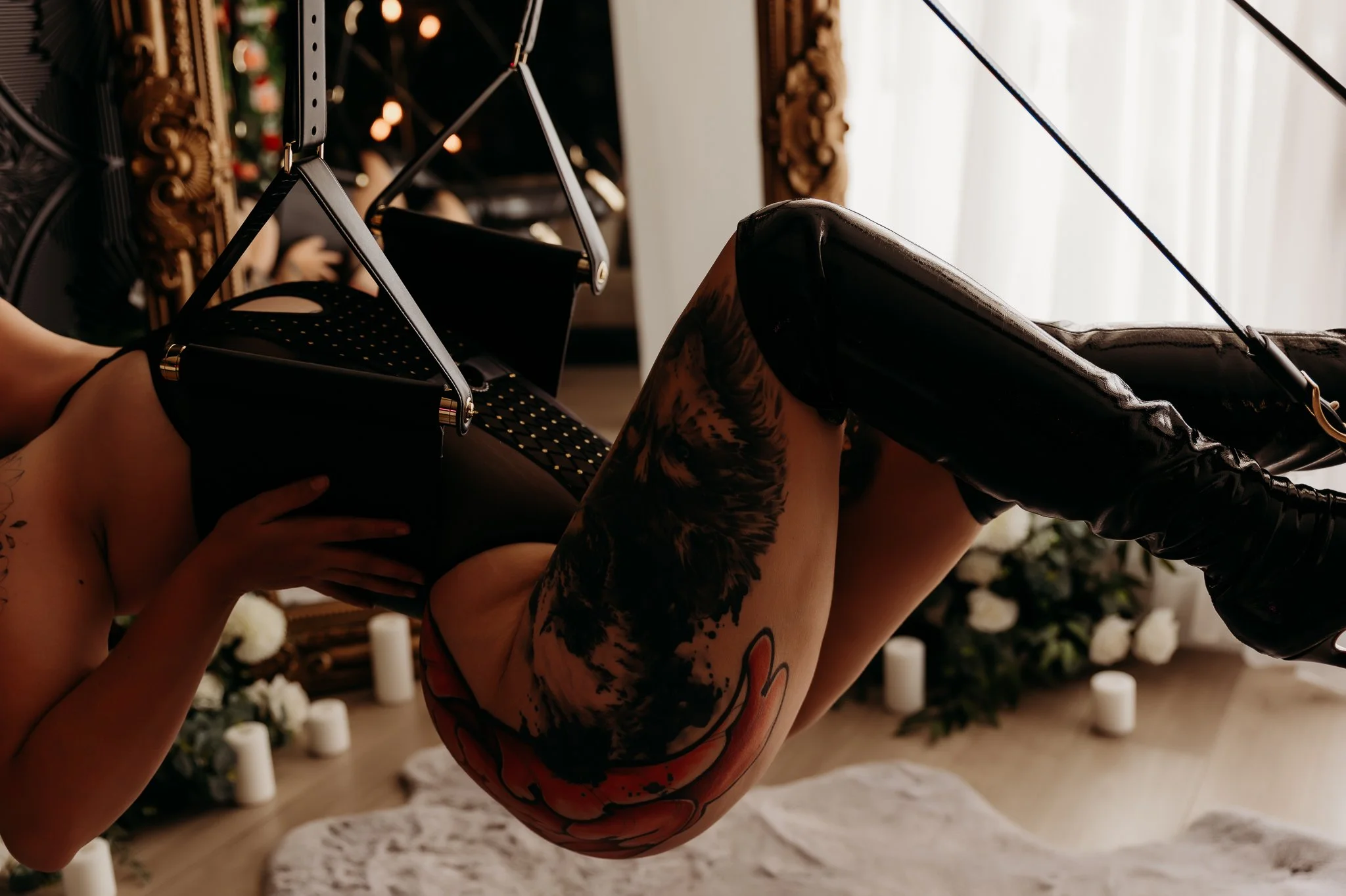 A tattooed woman lying on her back with her legs raised and resting against another person's knees, in a room with candlelit background and white candles on the floor.