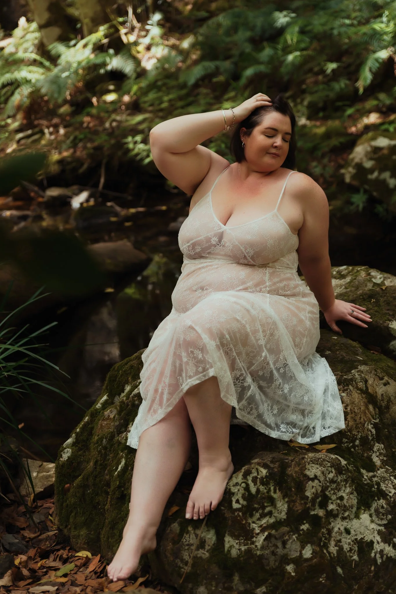 A woman in a white lace dress sitting on a mossy rock in a forest, with her eyes closed, touching her hair.