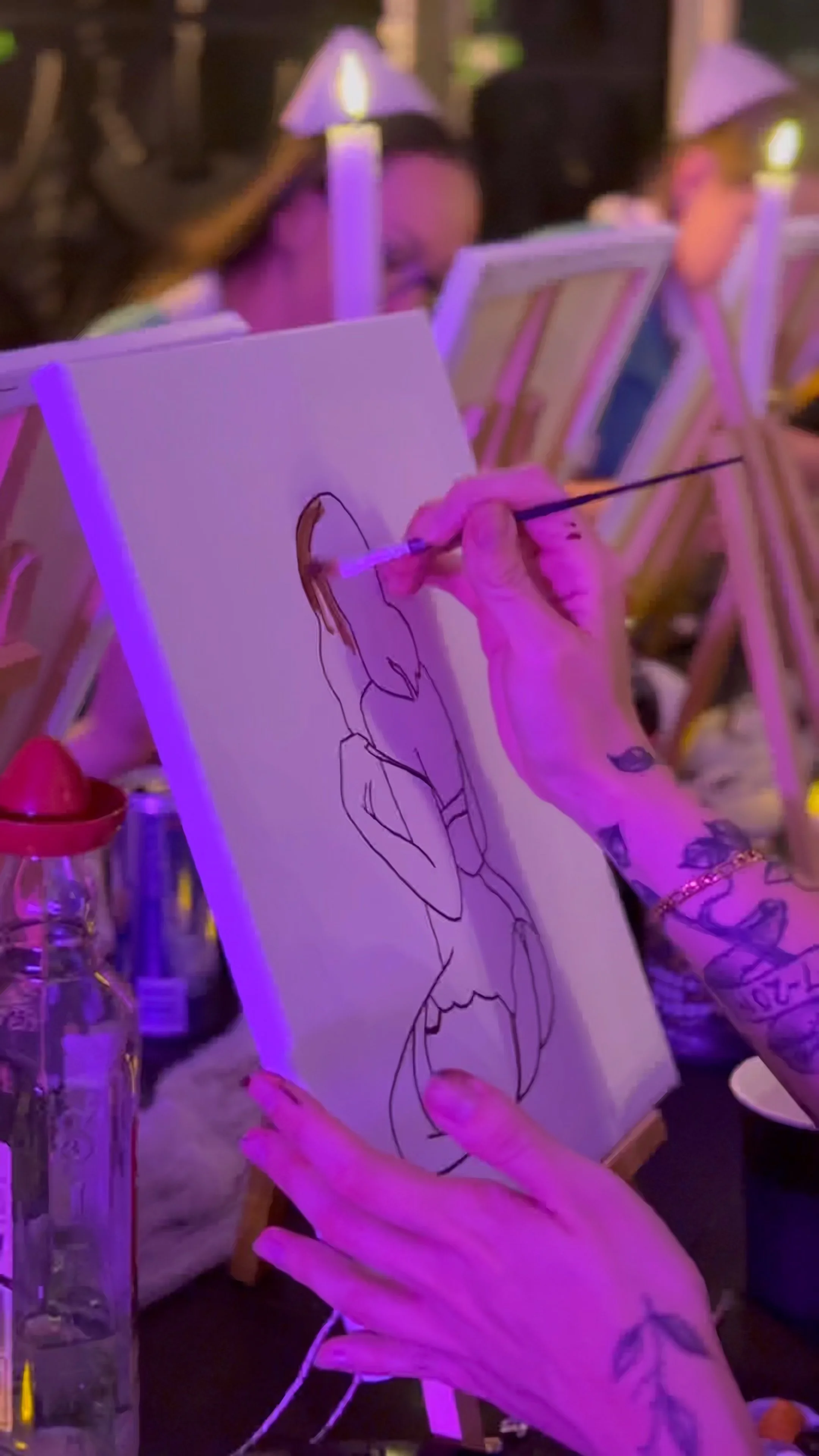 Person with tattoos painting on a canvas at a social event, with other people and canvases visible in the background, illuminated by purple lighting.