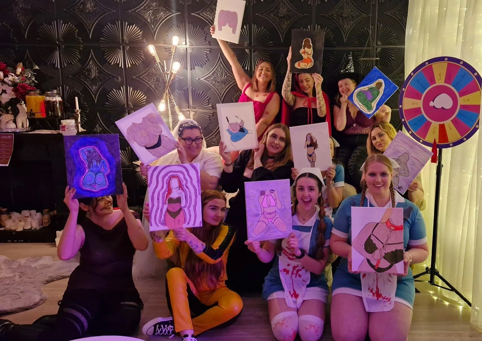 Group of women at a bachelorette party, holding colorful drawings of women in lingerie, with a decorated party room and a spinning wheel in the background.