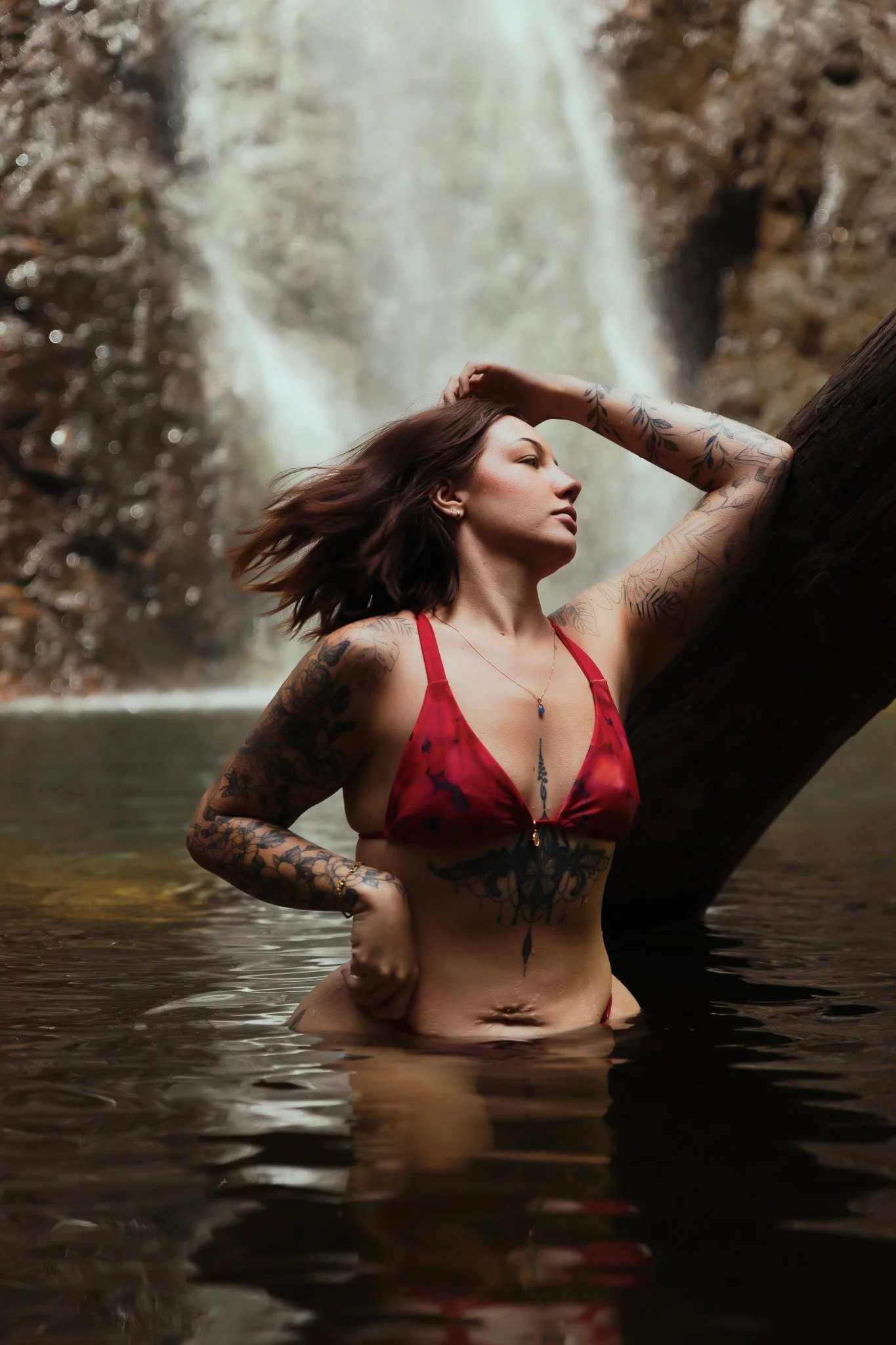 A woman in a red bikini with tattoos on her arms and stomach, standing waist-deep in a river, leaning against a large tree trunk with a waterfall in the background.
