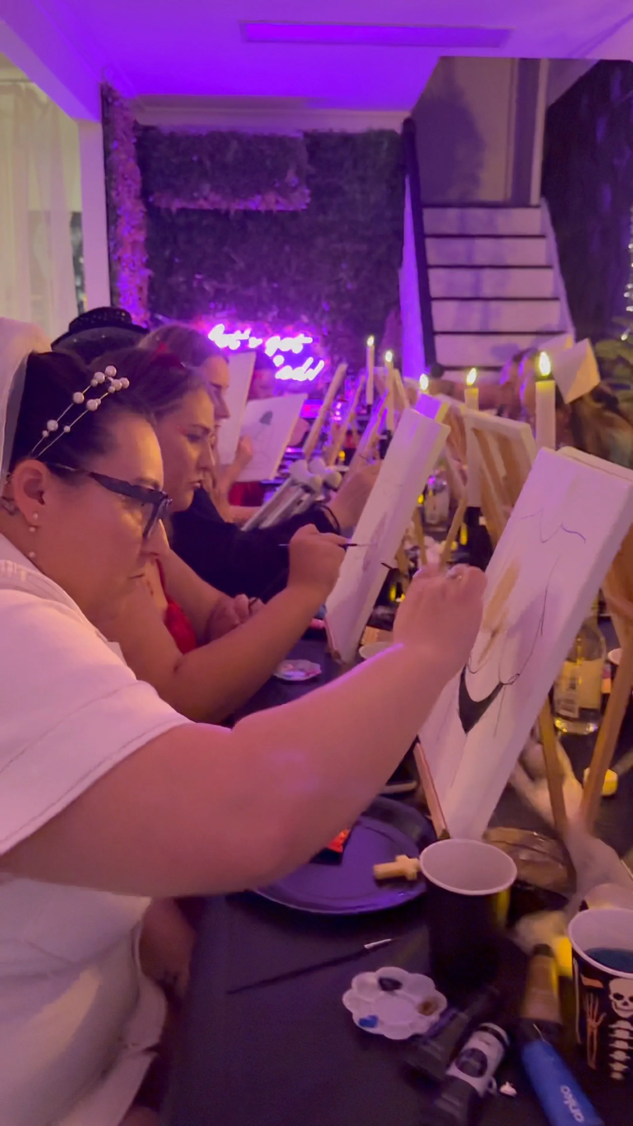 People sitting at tables painting on canvases, illuminated by candles, with a neon sign and purple lighting in a cozy indoor setting.