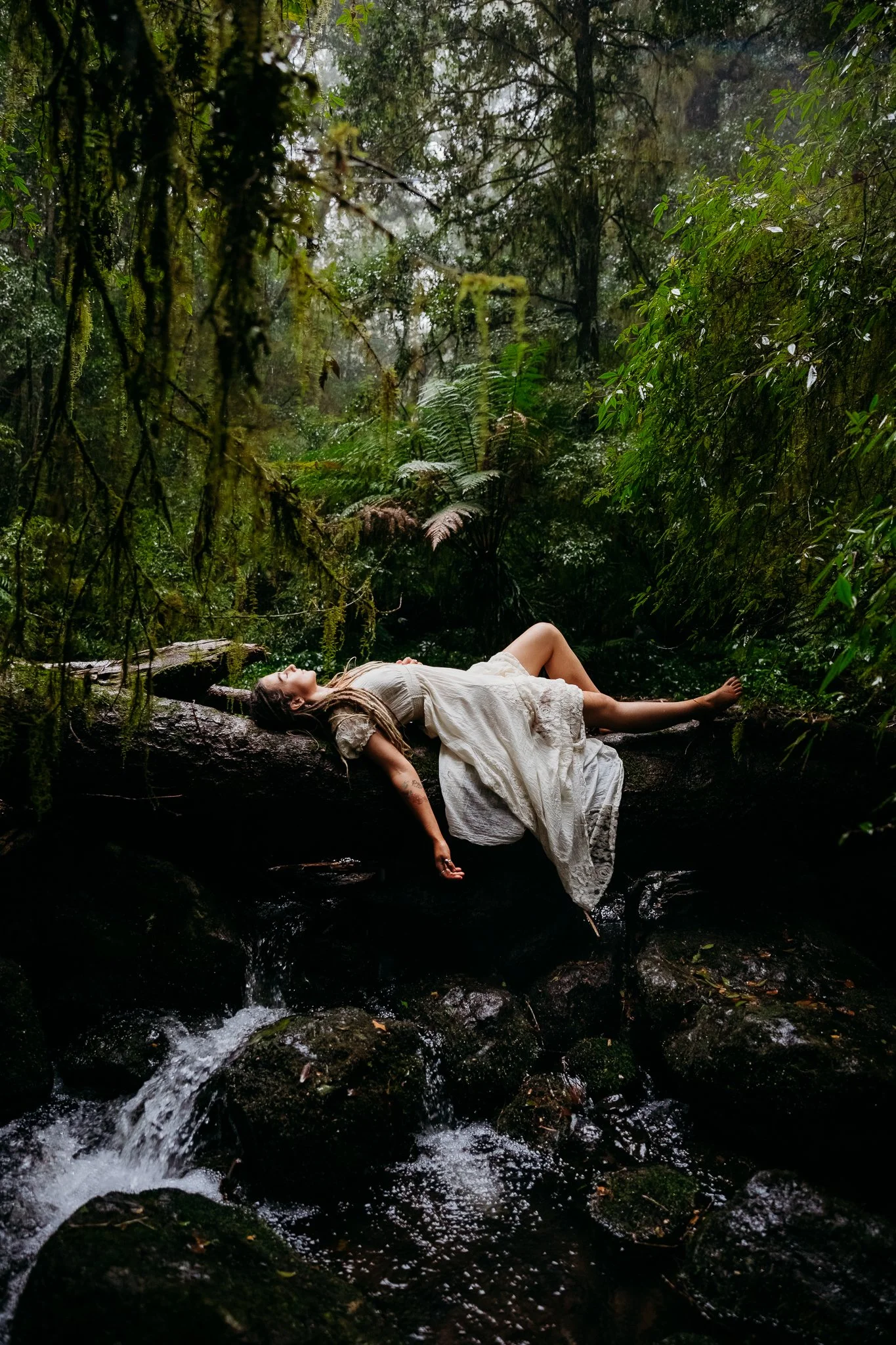 A woman in a long beige dress laying on a fallen tree in a lush, green forest by a small stream.