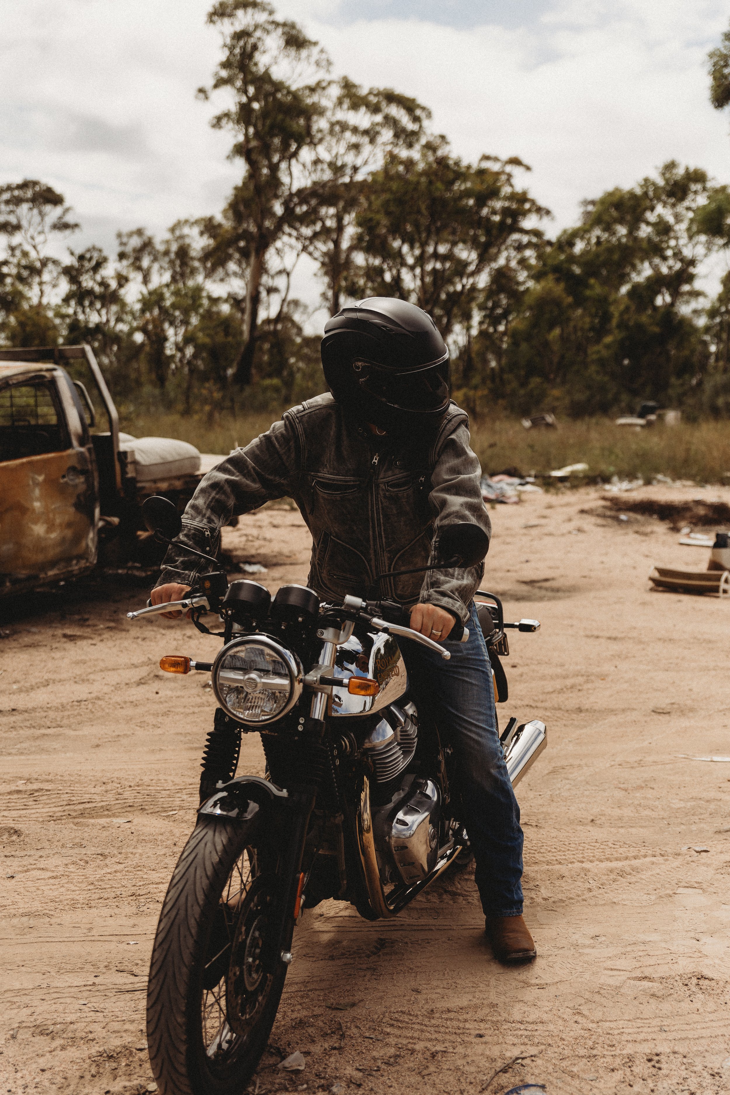 Person in a black helmet and leather jacket riding a motorcycle on a dirt path with trees in the background.