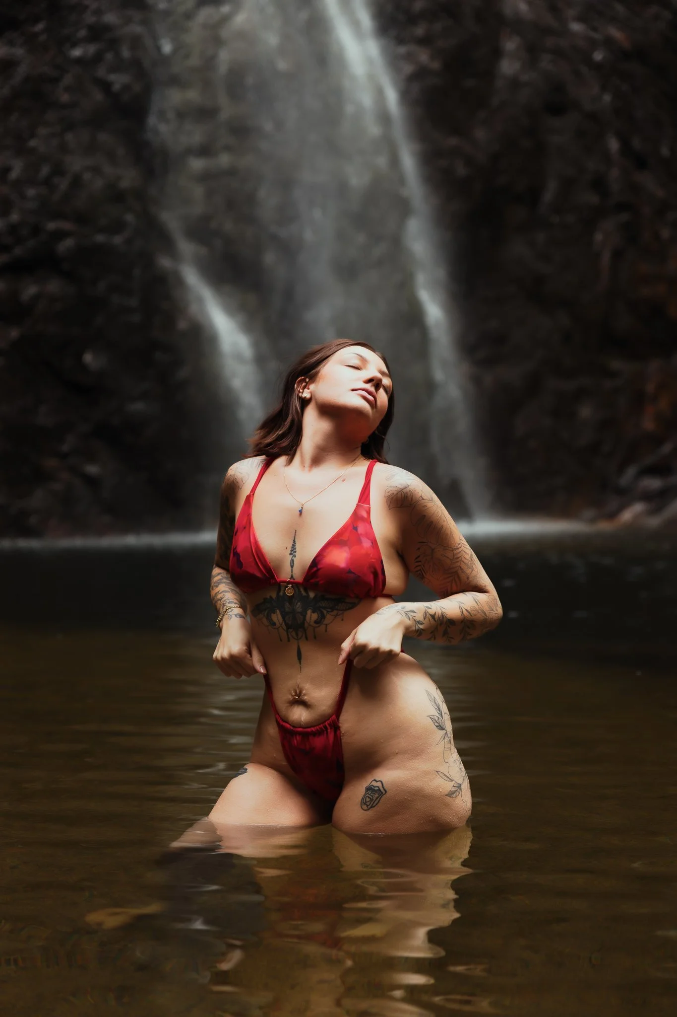 A woman with tattoos in a red bikini standing in a body of water with a waterfall in the background.