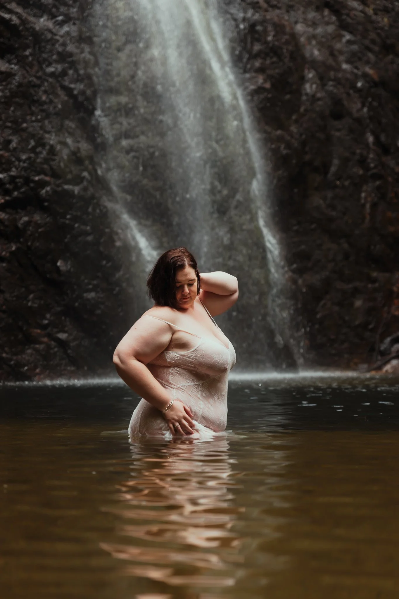 A woman standing in a body of water with a waterfall cascading behind her in a natural setting.