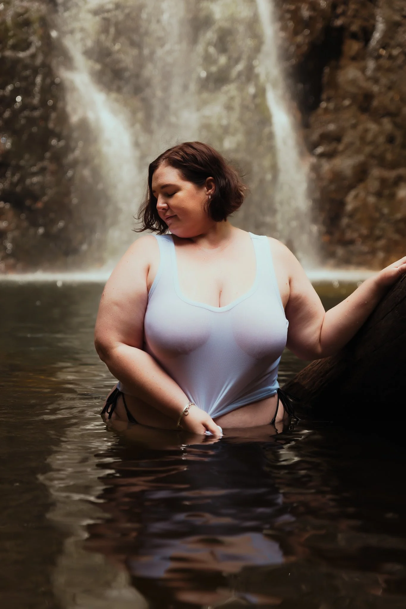 Woman standing in water near a waterfall, wearing a white tank top and black bikini bottoms, with brown hair and a bracelet on her wrist.