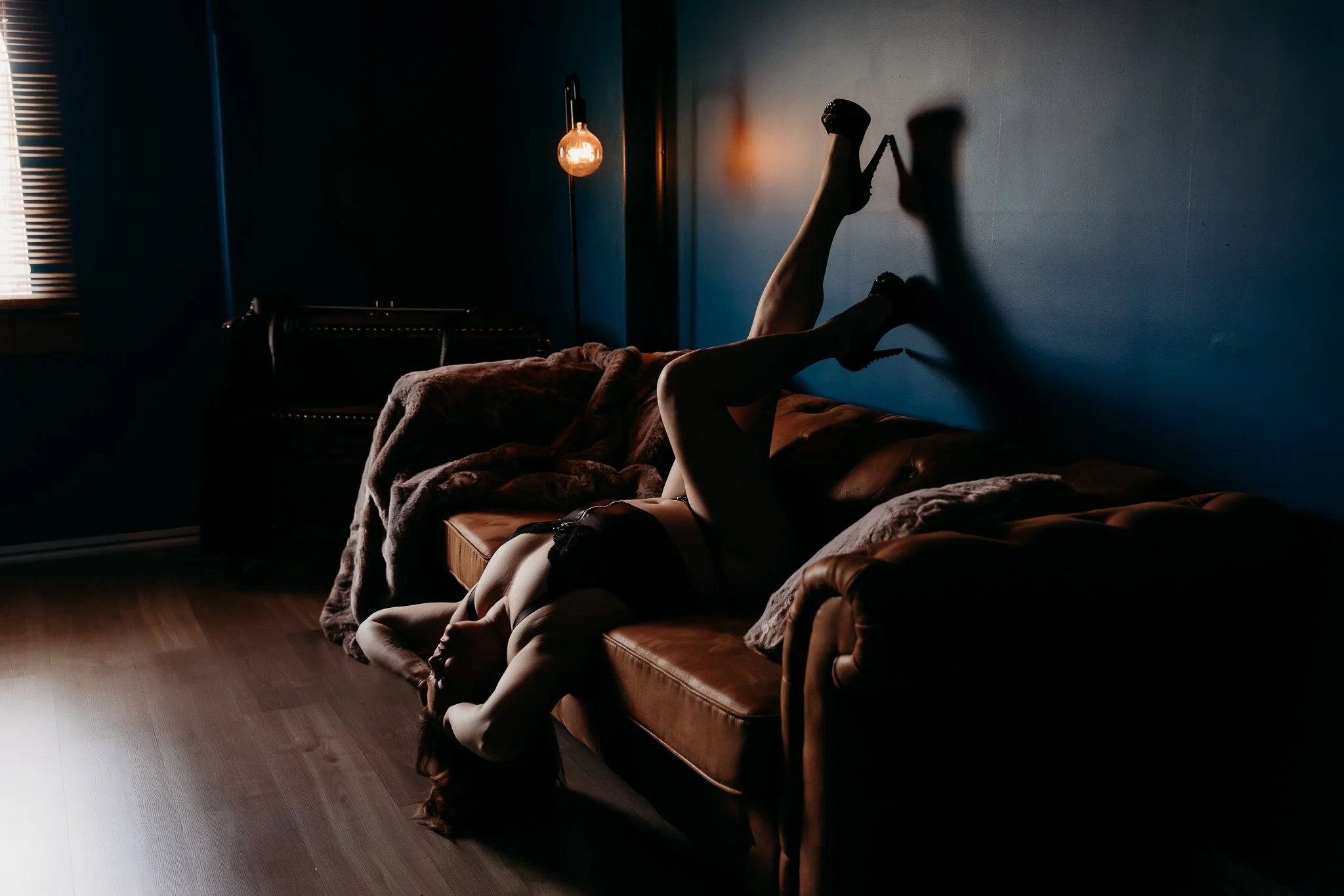 A woman in lingerie lying upside down on a sofa with her legs against the wall in a dimly lit room.