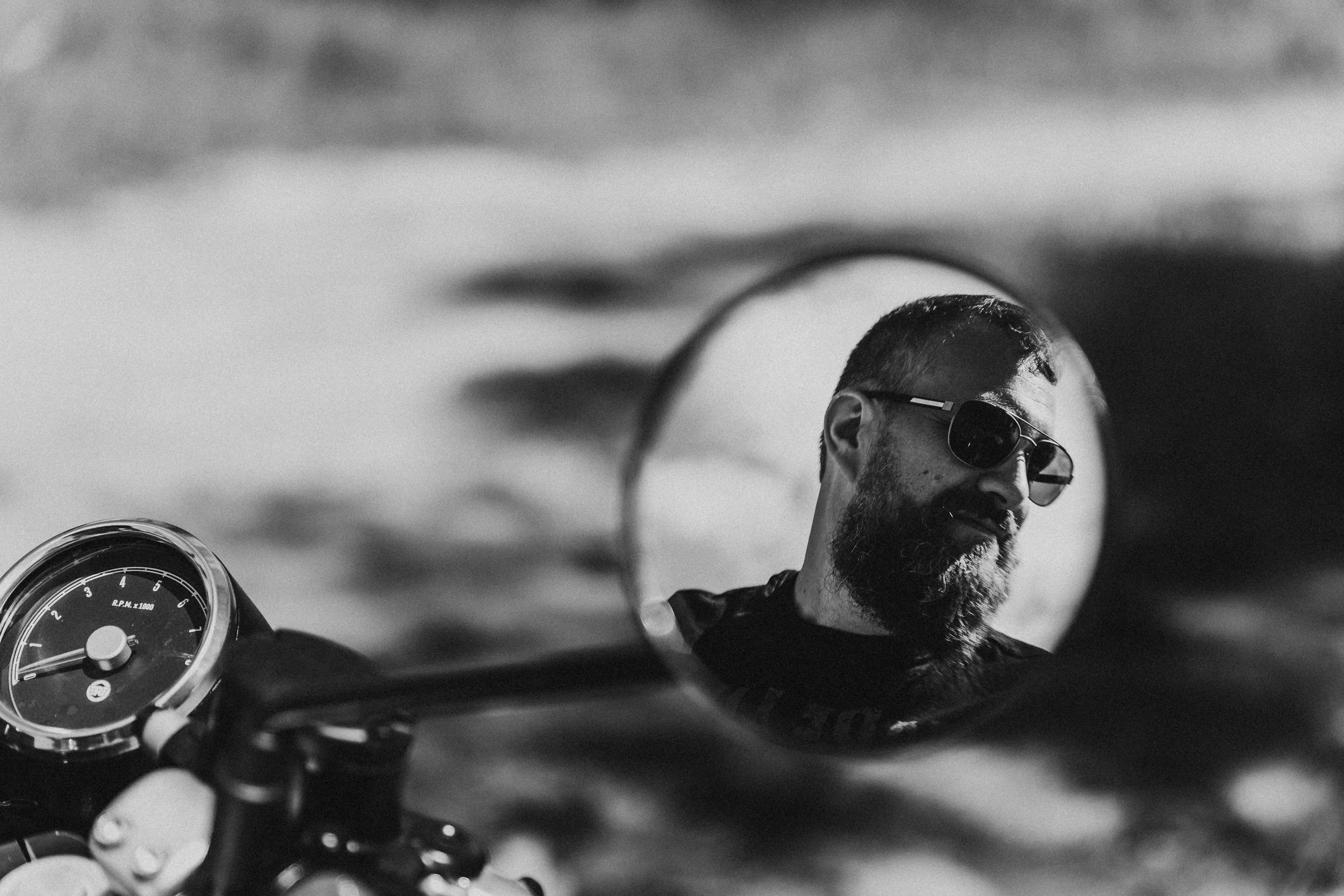 A reflection of a man with sunglasses and a beard on a motorcycle's side mirror, with the motorcycle's speedometer visible in the foreground.