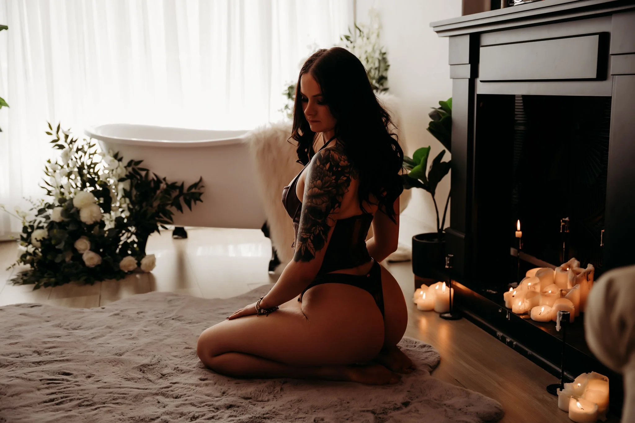A woman in black lingerie kneeling on a plush rug by a fireplace with candles, in a softly lit room with a bathtub and flowers in the background.