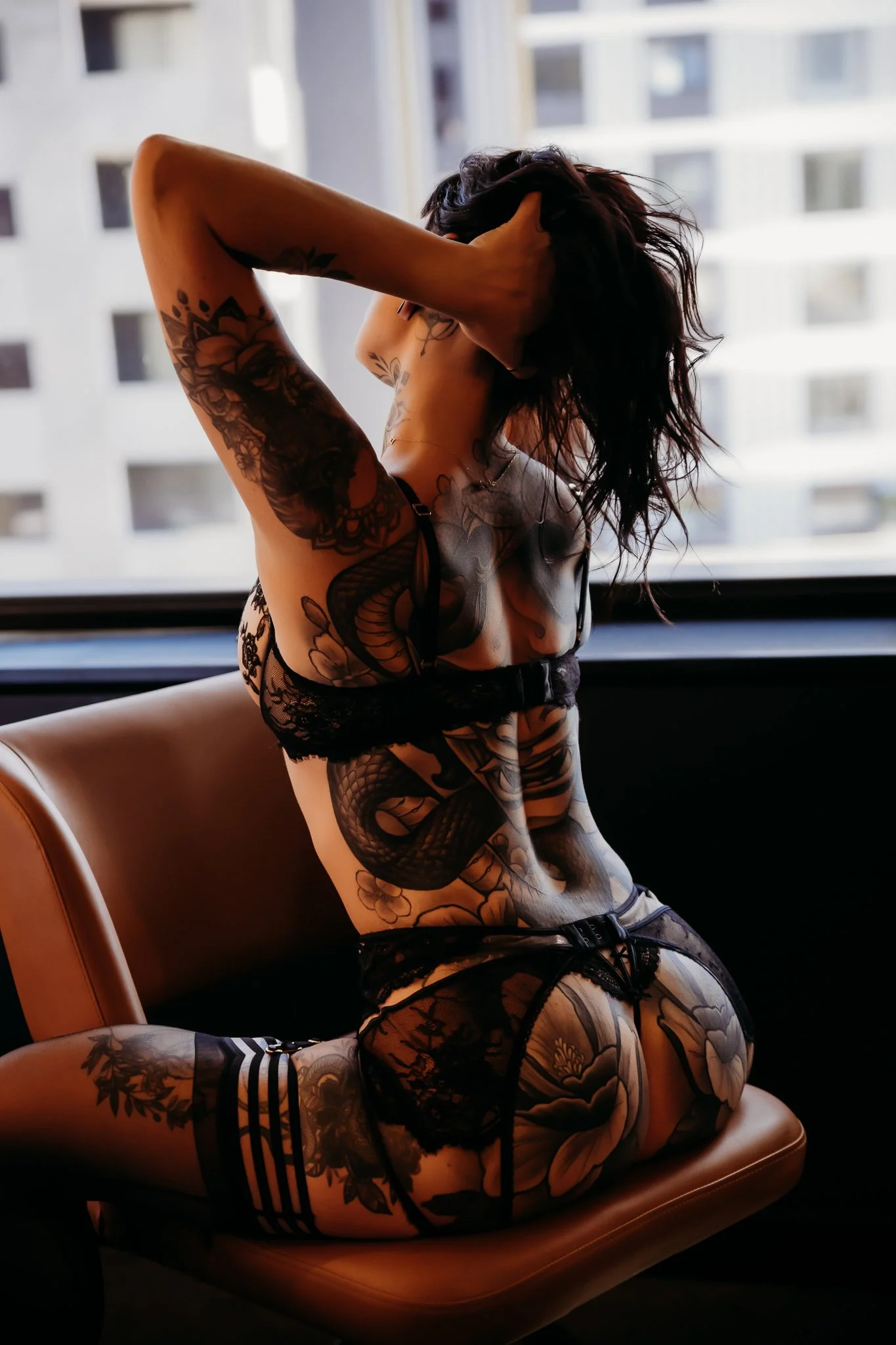 A woman with elaborate tattoos wearing lingerie, sitting on a chair near a window with city buildings in the background.