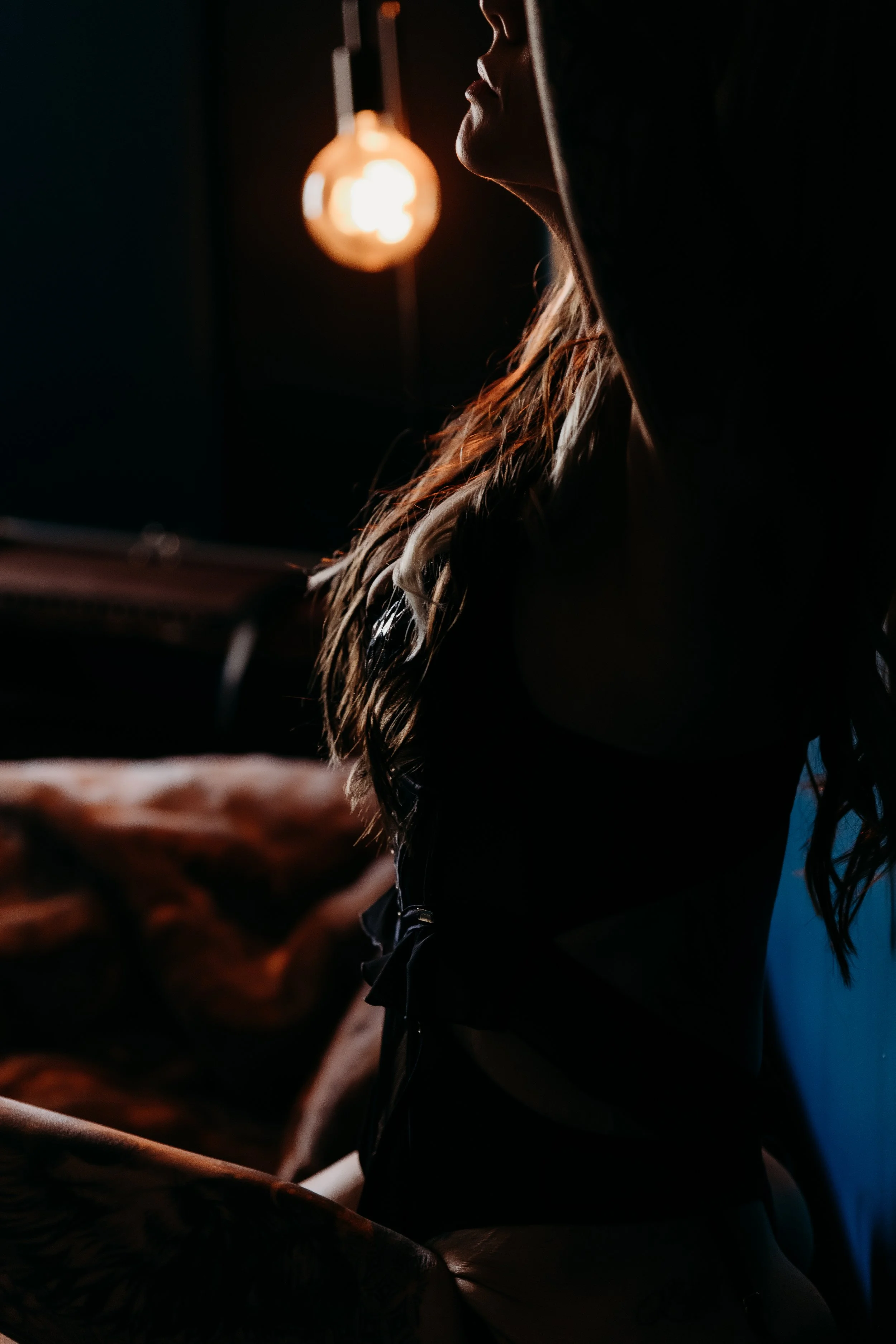 Side view of a woman with long wavy hair sitting in a dimly lit room, illuminated by an orange hanging light bulb.