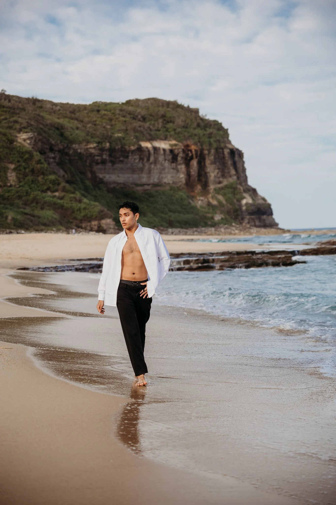 A man walking along a sandy beach near the ocean with greenery-covered cliffs in the background.