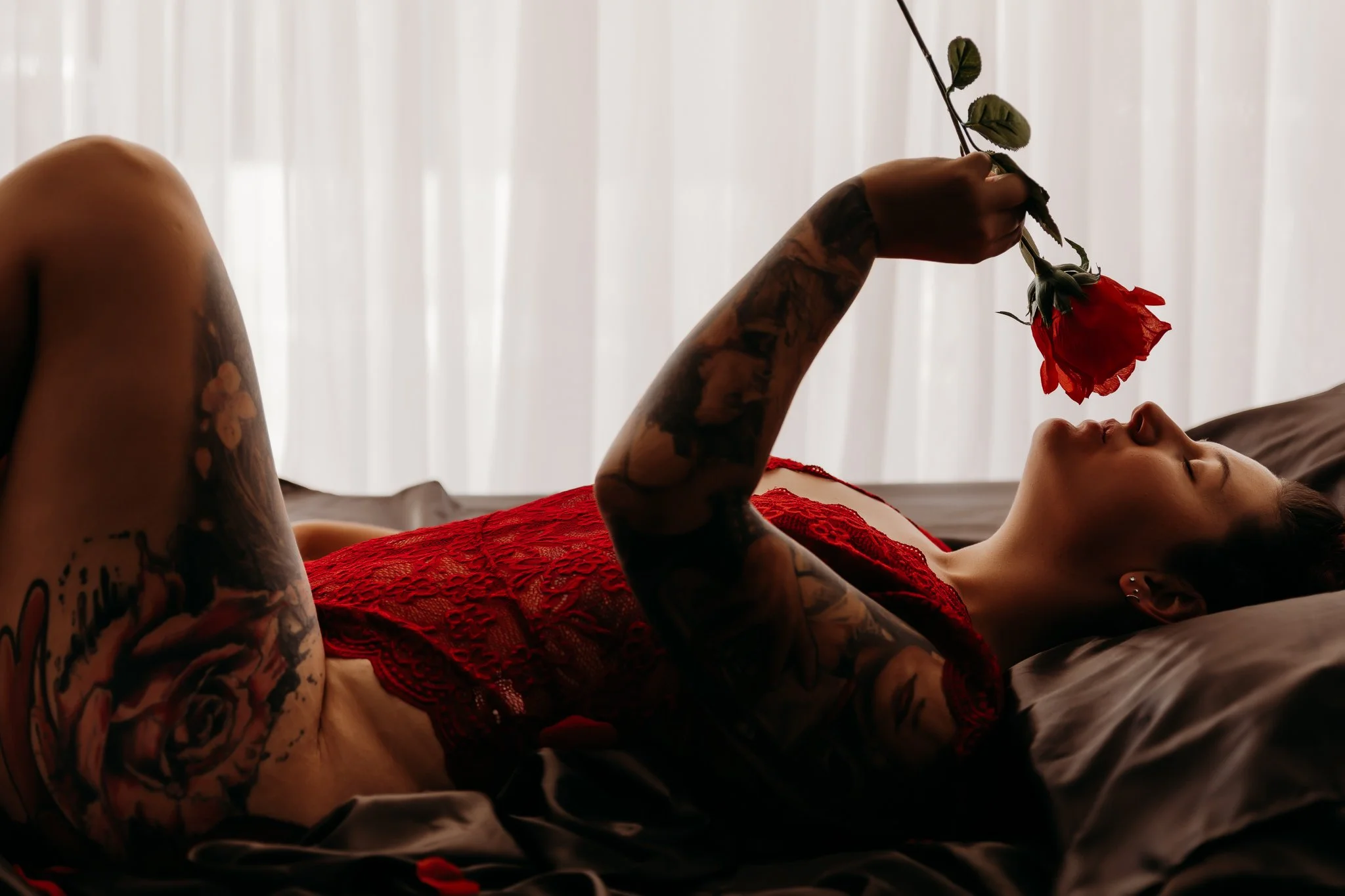 A woman with tattoos lying on a bed, holding a red rose, wearing a red lace dress, with her eyes closed.