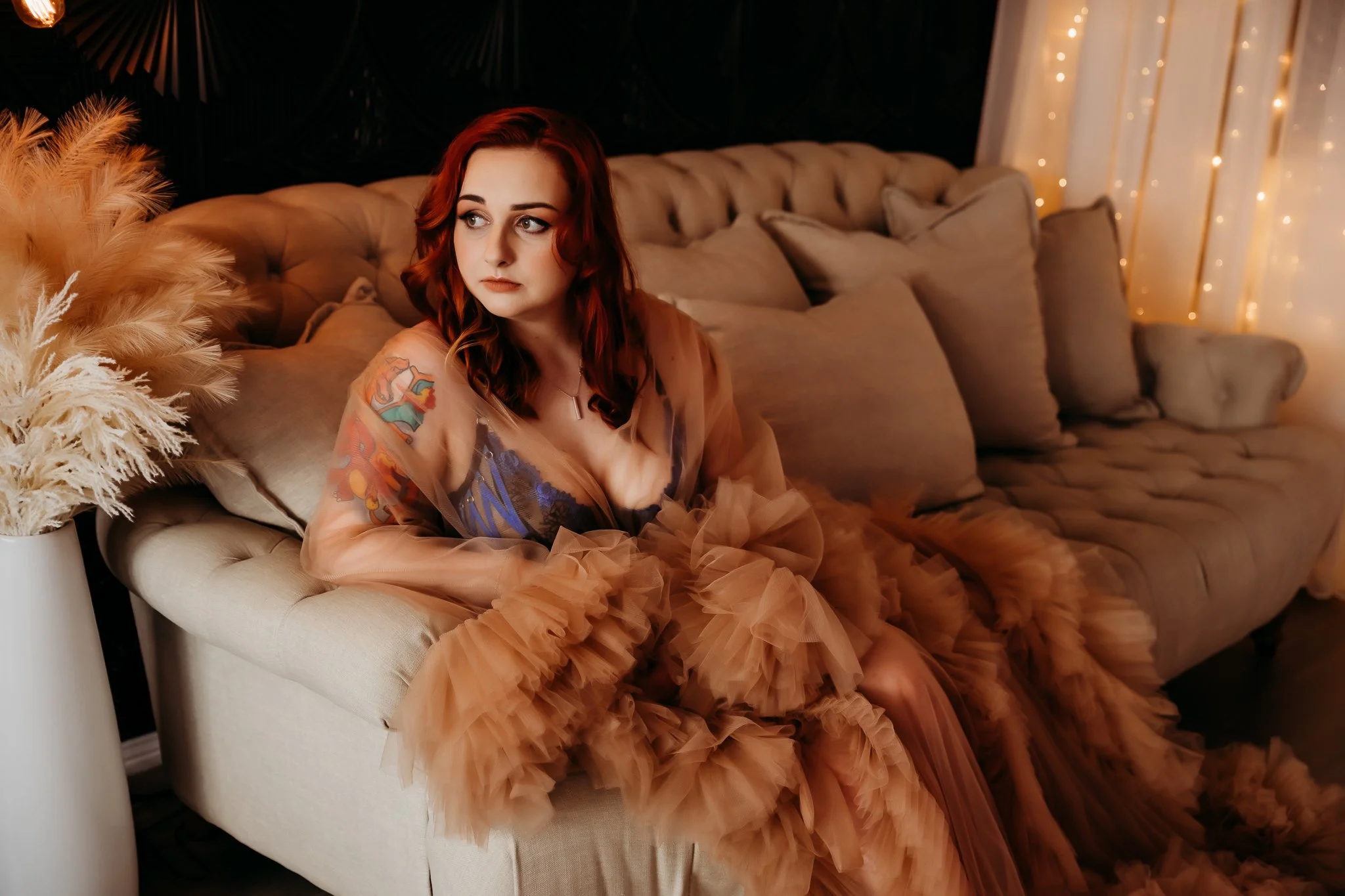 A woman with red hair and tattoos, wearing a brown tulle dress, sitting on a beige tufted sofa in a decorated room with fairy lights.