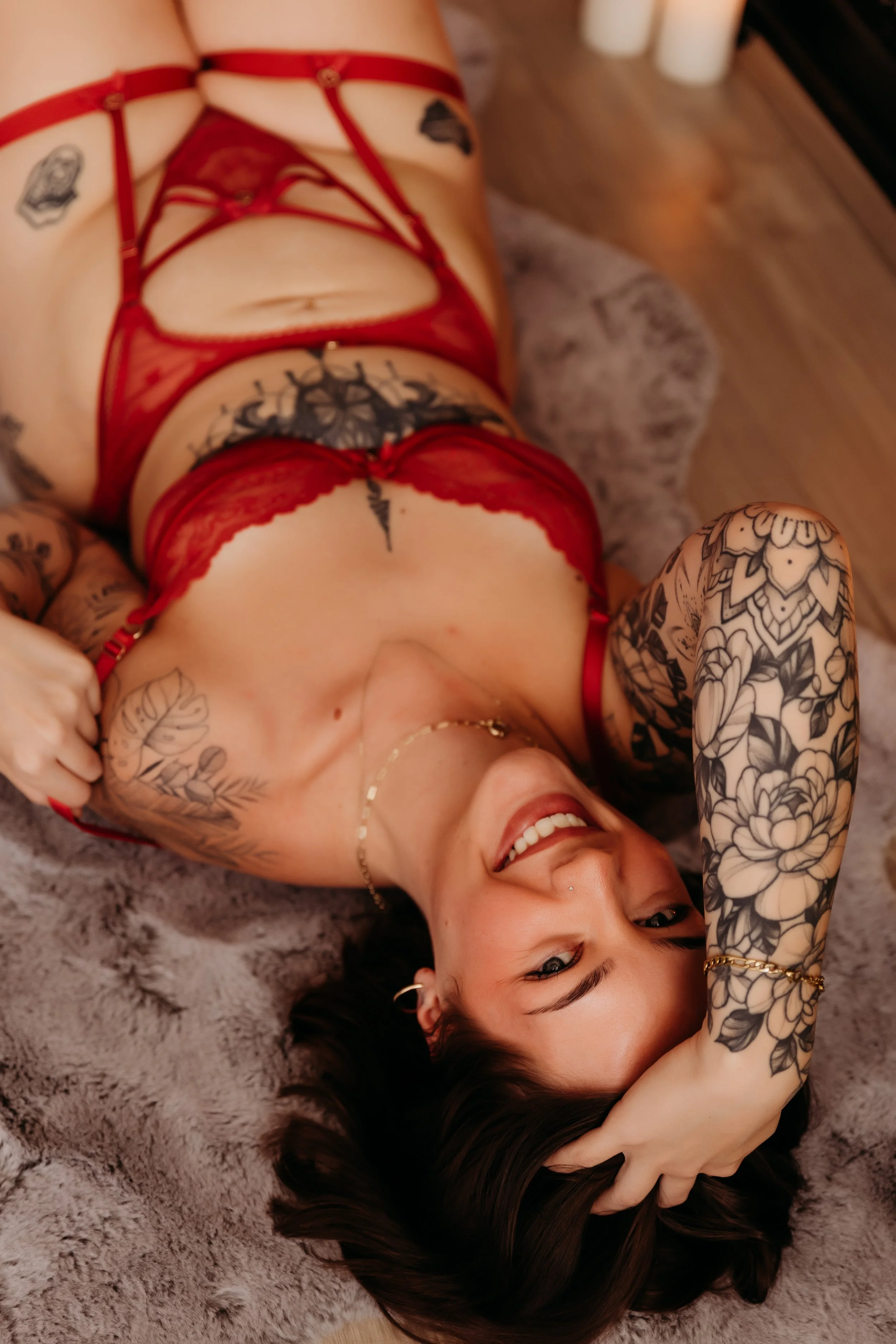 A woman lying on a fuzzy rug, wearing red lingerie, with tattoos on her arm and chest, smiling and looking at the camera, with her hand on her forehead and dark hair spread out on the rug.