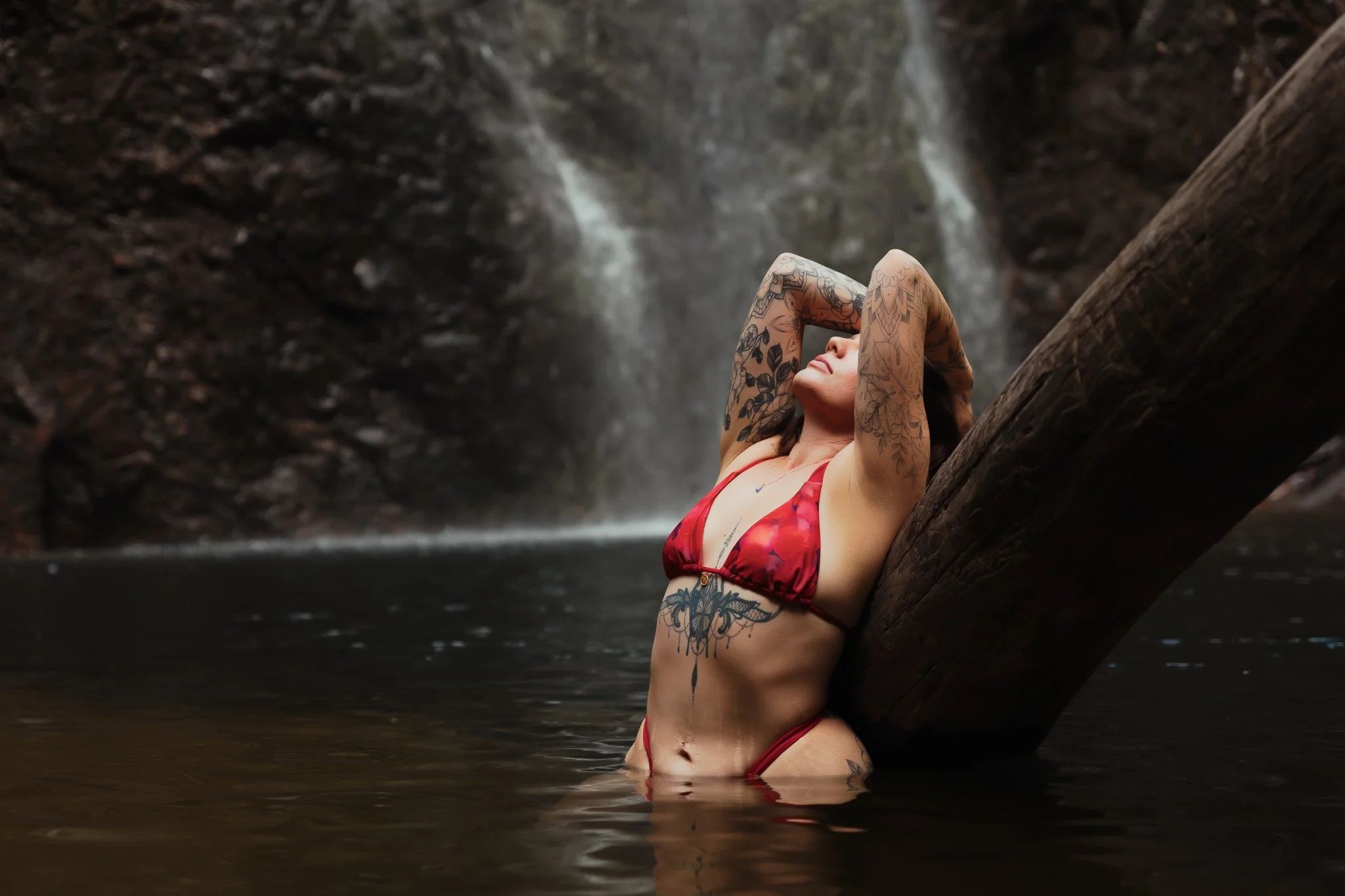 Woman with tattoos in red bikini leaning back against a fallen log in water near a waterfall