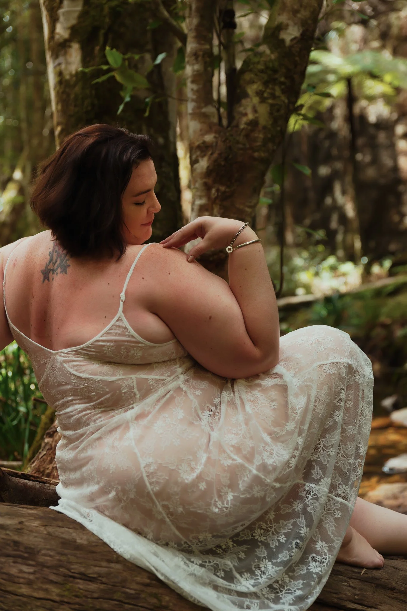 A woman with dark hair, tattoos, and wearing a white lace dress sitting on a log outdoors near a tree in a forested area.