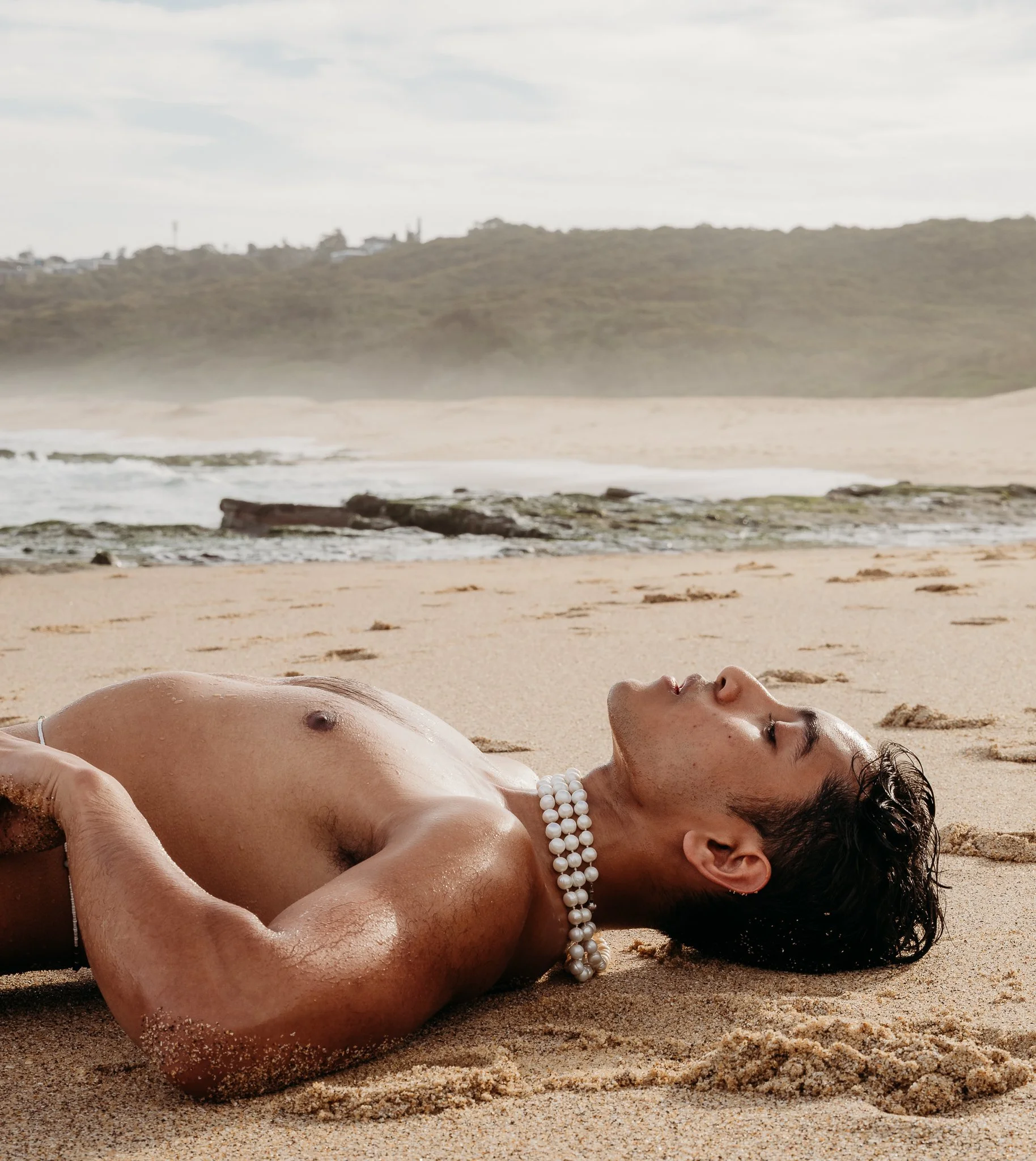 A person lying on a sandy beach with their eyes closed, wearing pearl necklaces, with ocean waves and a cloudy sky in the background.