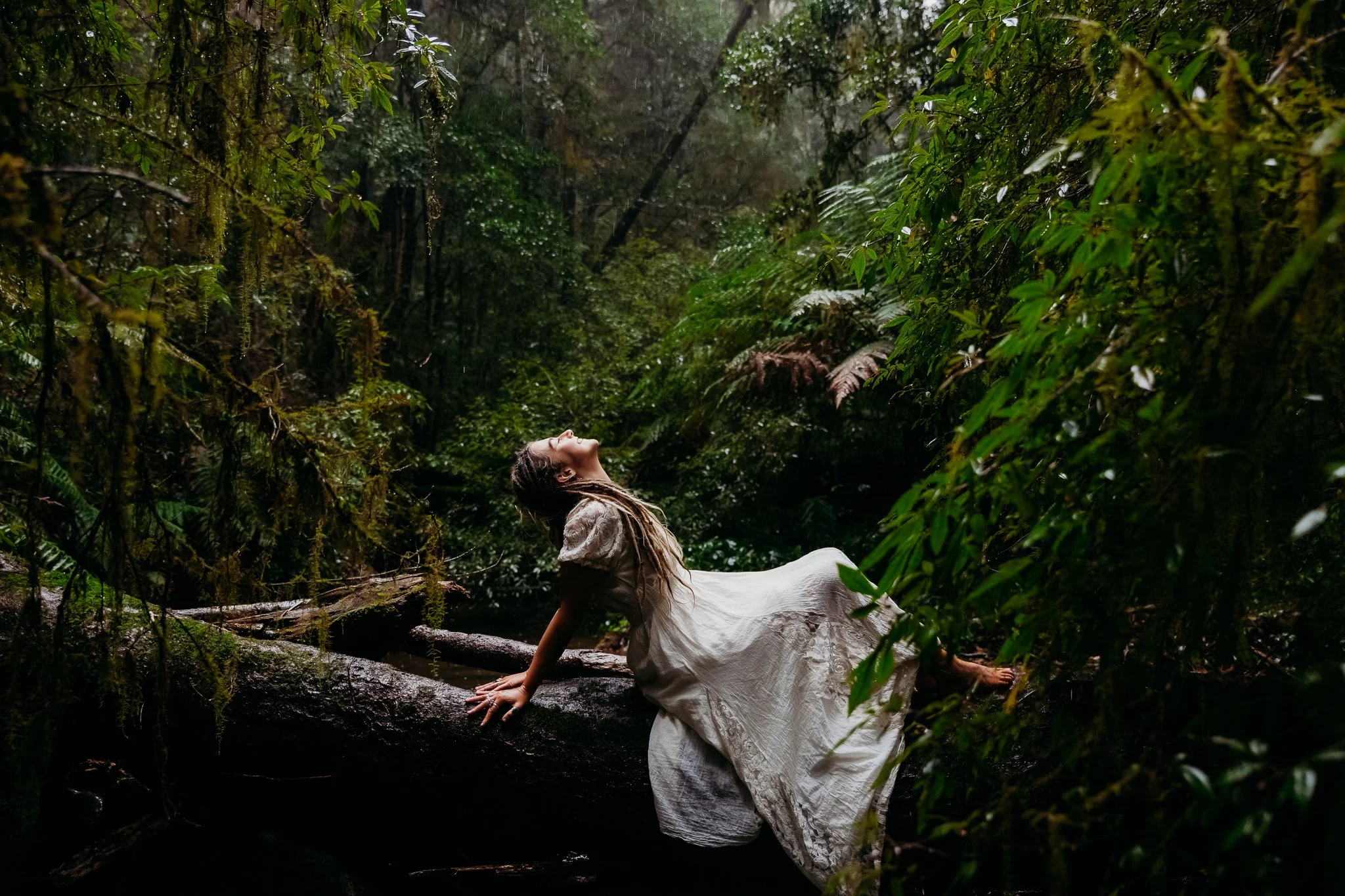 A woman in a white dress reclines on a fallen tree in a dense, green jungle, with her head tilted back and eyes closed, surrounded by lush foliage and moss-covered branches.