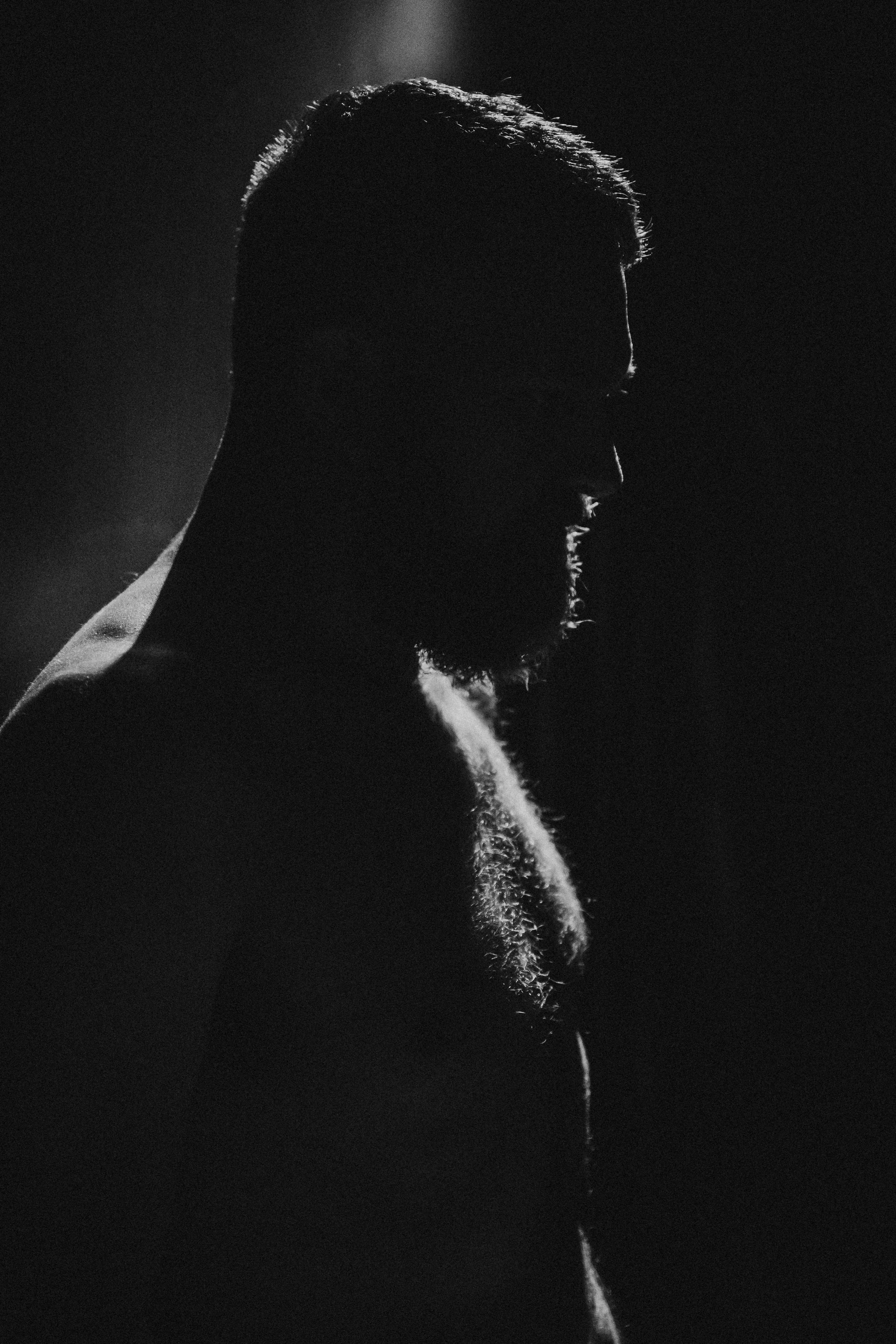Black and white silhouette of a man with a beard, looking downward, with a light source behind him highlighting the edges of his head and shoulders.