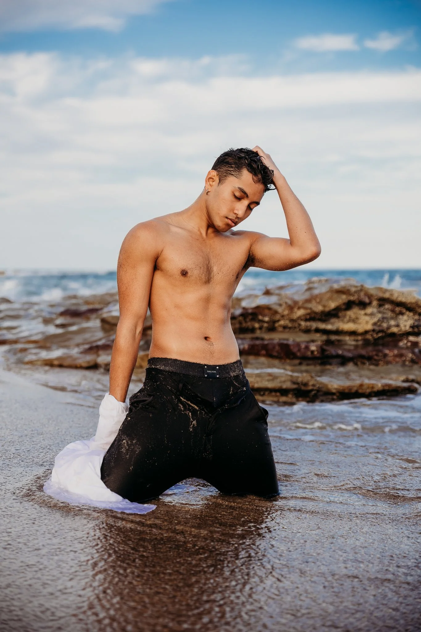 A shirtless man with wet hair kneeling in shallow ocean water with rocks in the background, holding a white shirt in his left hand, looking down and touching his head with his right hand.