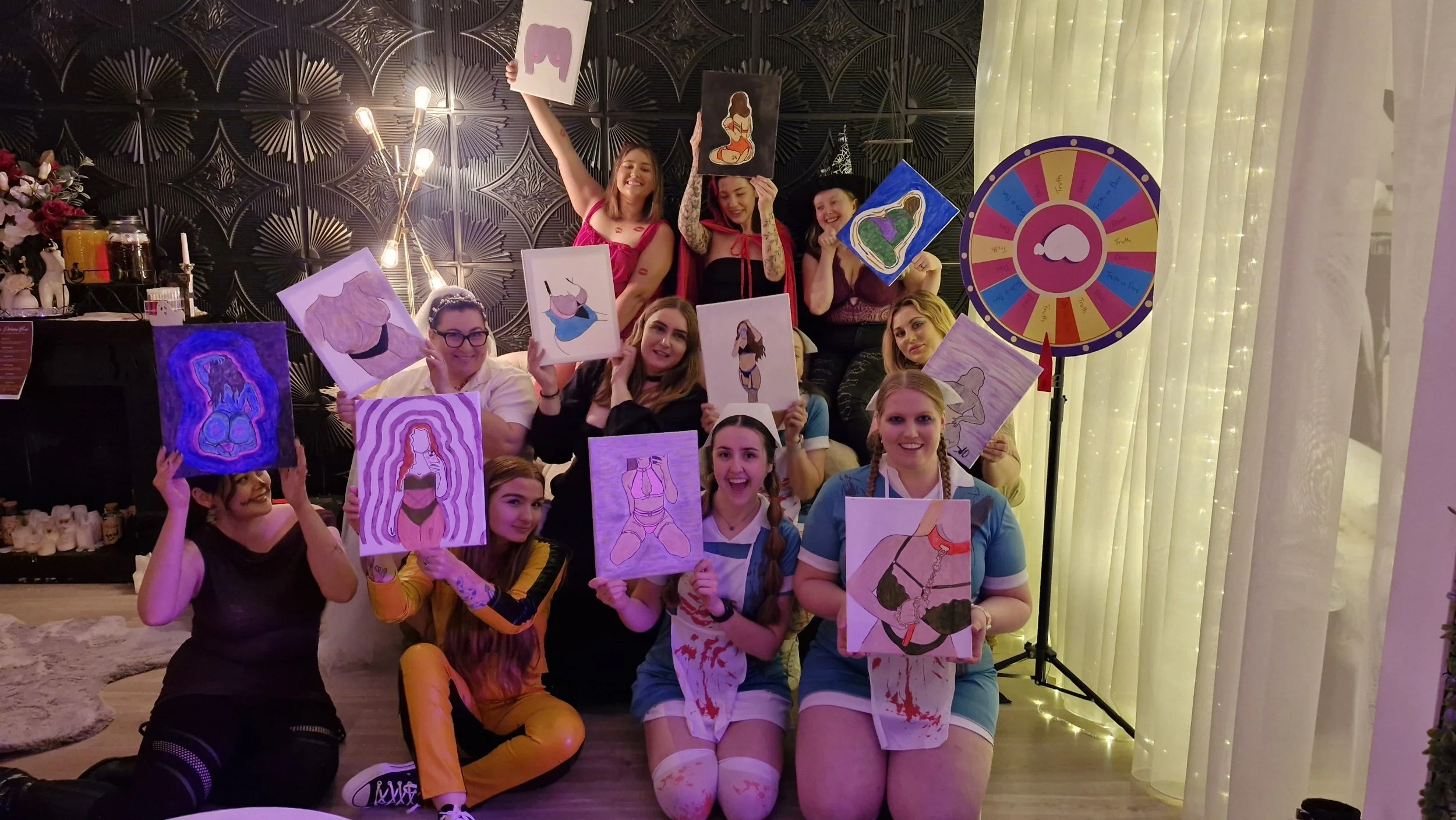 A group of young women celebrating at a party, holding up provocative drawings of female figures, with a colorful spinning wheel in the background and festive decorations.