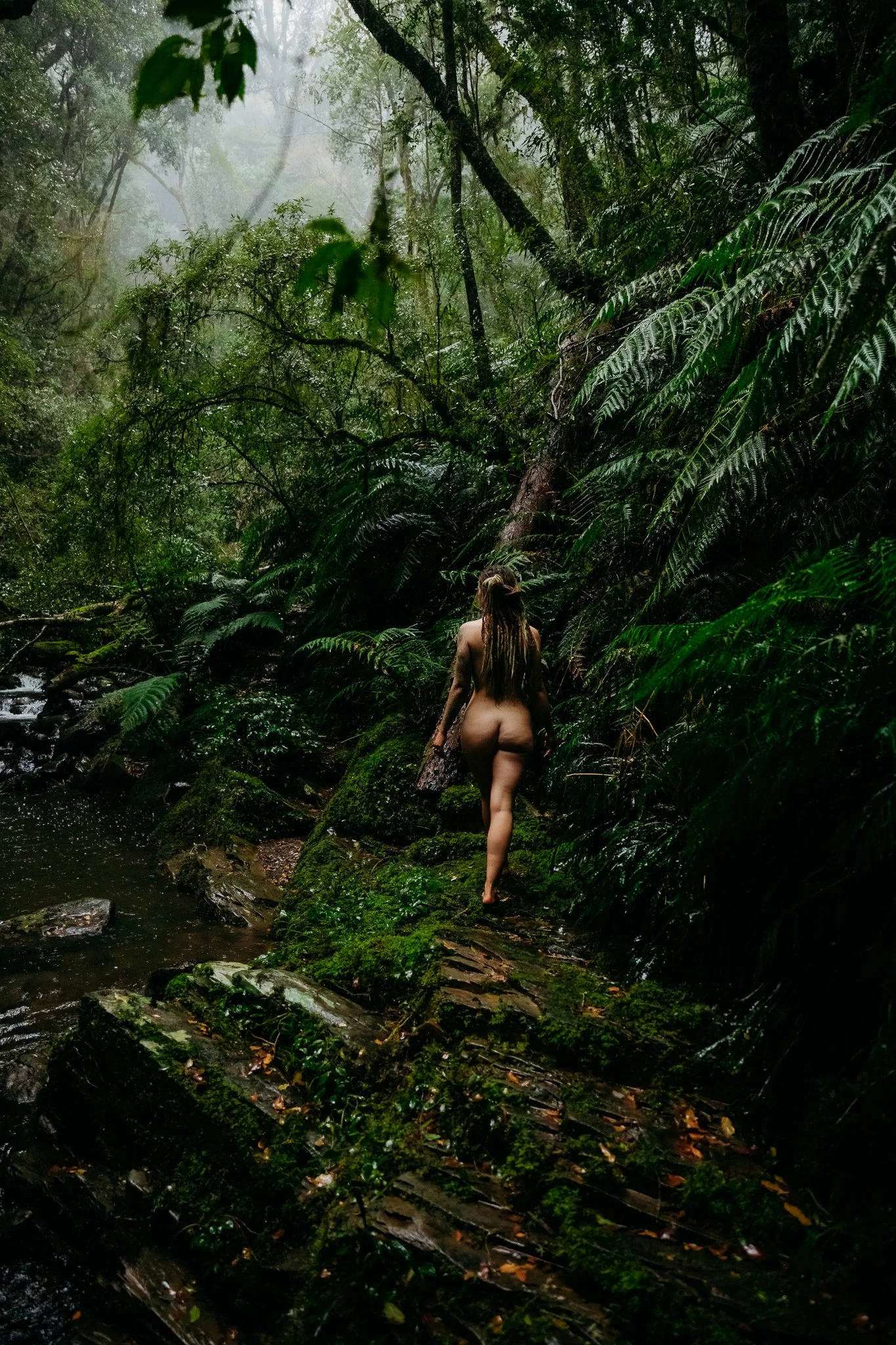 A woman with long hair walking along a mossy trail in a lush, green rainforest near a stream.