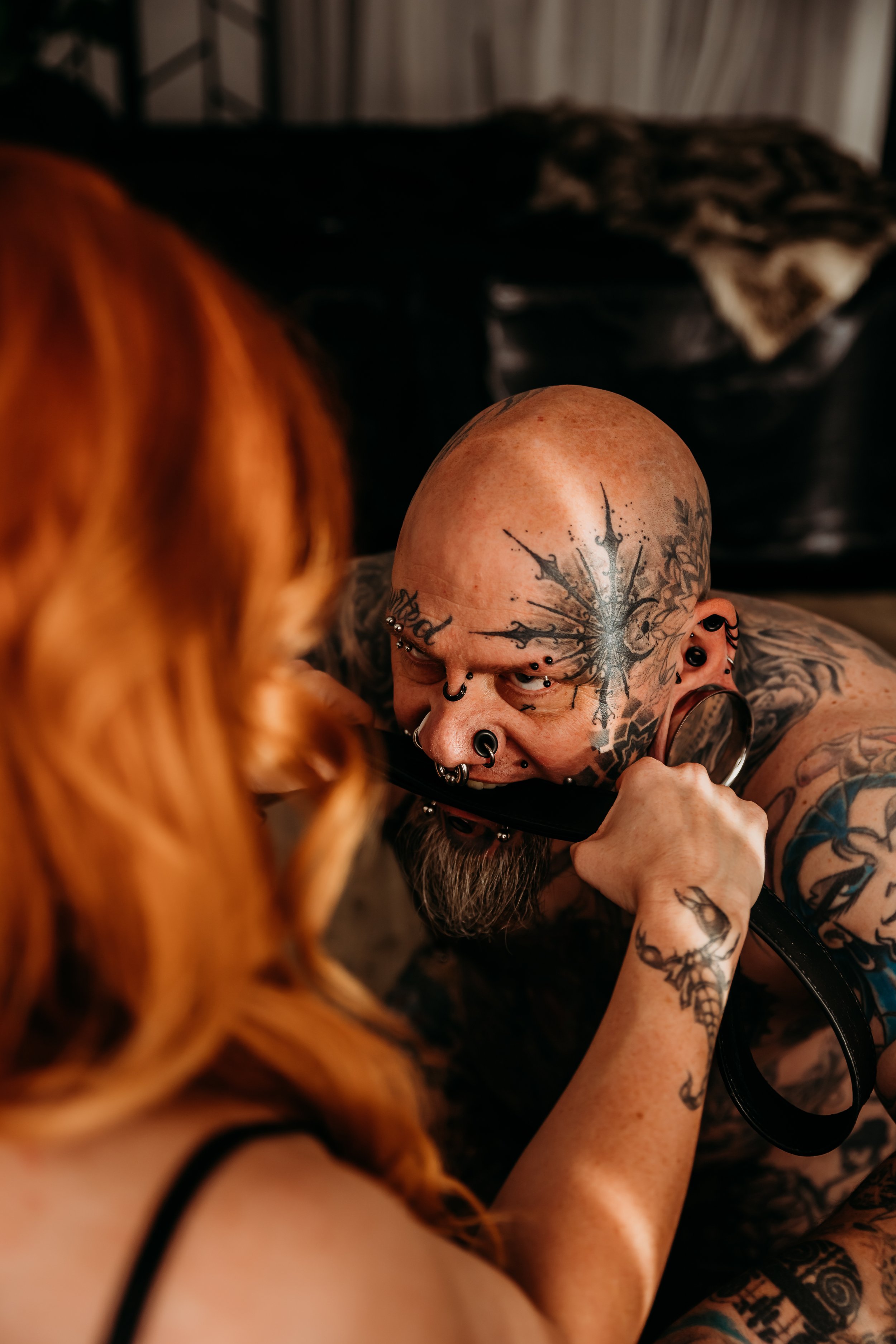 A tattooed bald man with multiple facial piercings holding a belt in his mouth, interacting closely with a woman with red hair in a dimly lit room.