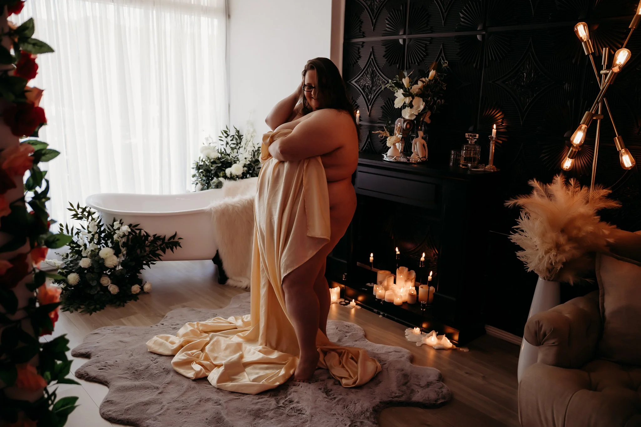 A woman standing on a rug with long beige satin fabric draped over her, in a dimly lit room with candles, floral arrangements, and a clawfoot bathtub in the background.