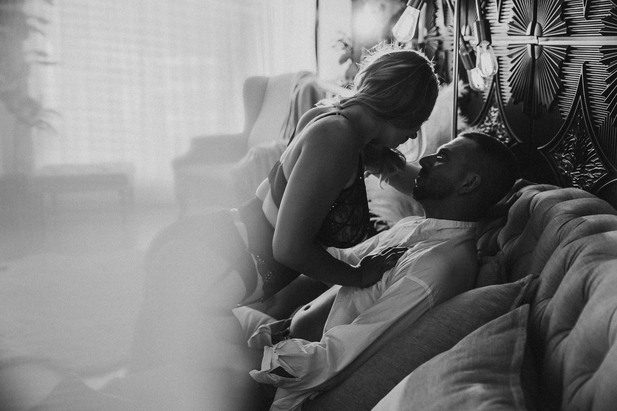 A black-and-white photo of a woman and a man on a bed in an intimate moment, with the woman leaning over the man who is lying down, both facing each other.