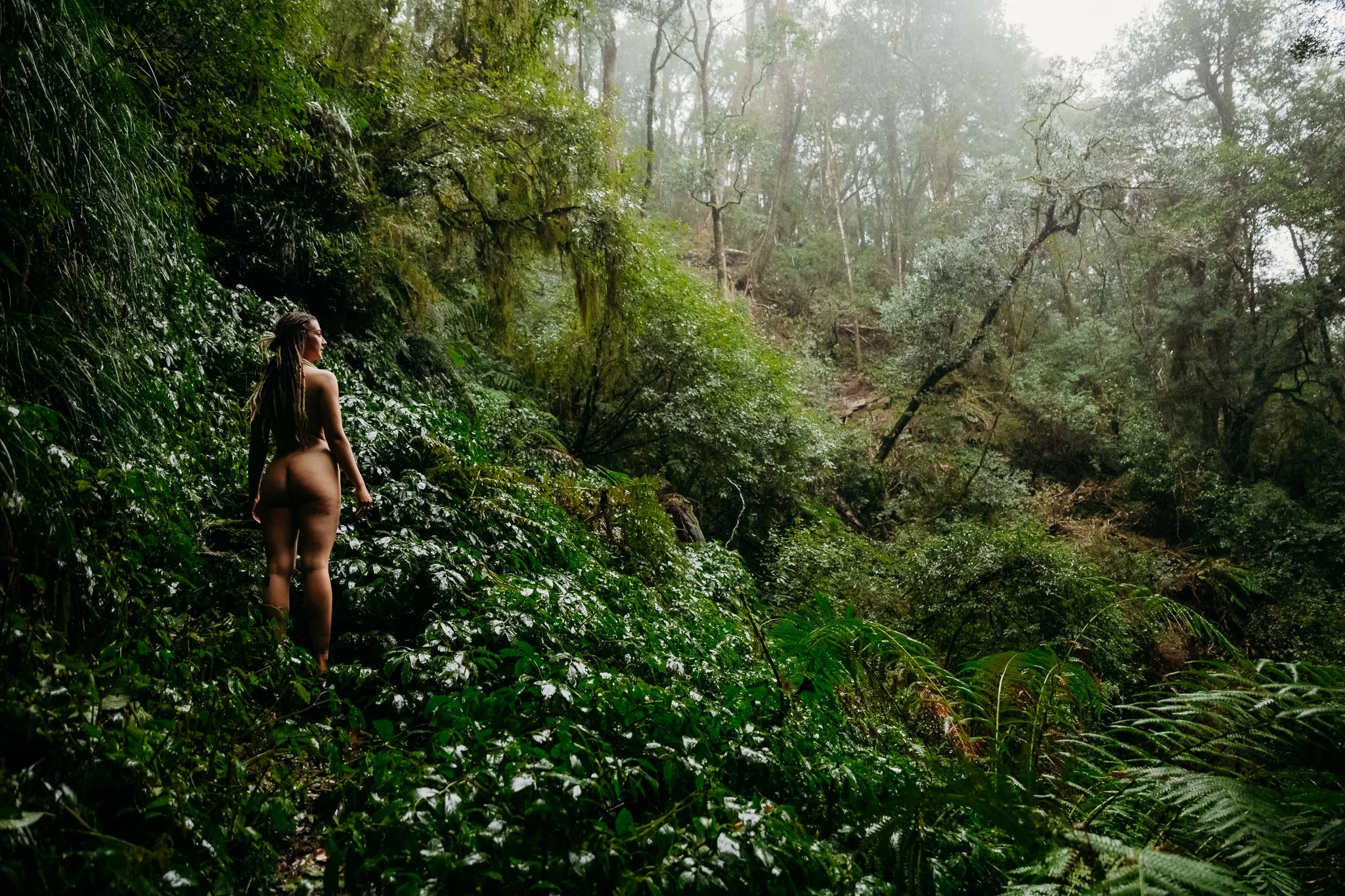 A woman with long hair standing nude in a lush green forest, surrounded by dense foliage and trees.