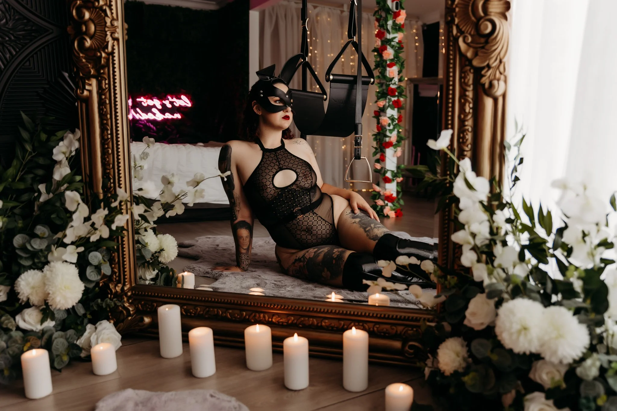 A woman dressed in black lingerie and a mask, sitting on the floor in front of a large ornate mirror surrounded by white flowers and candles, with fairy lights and floral decorations in the background.