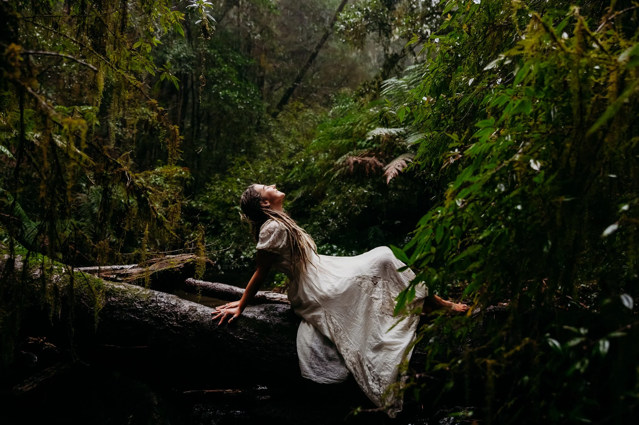 A woman in a white dress sitting on a log in a lush green forest, looking up with her head tilted back.