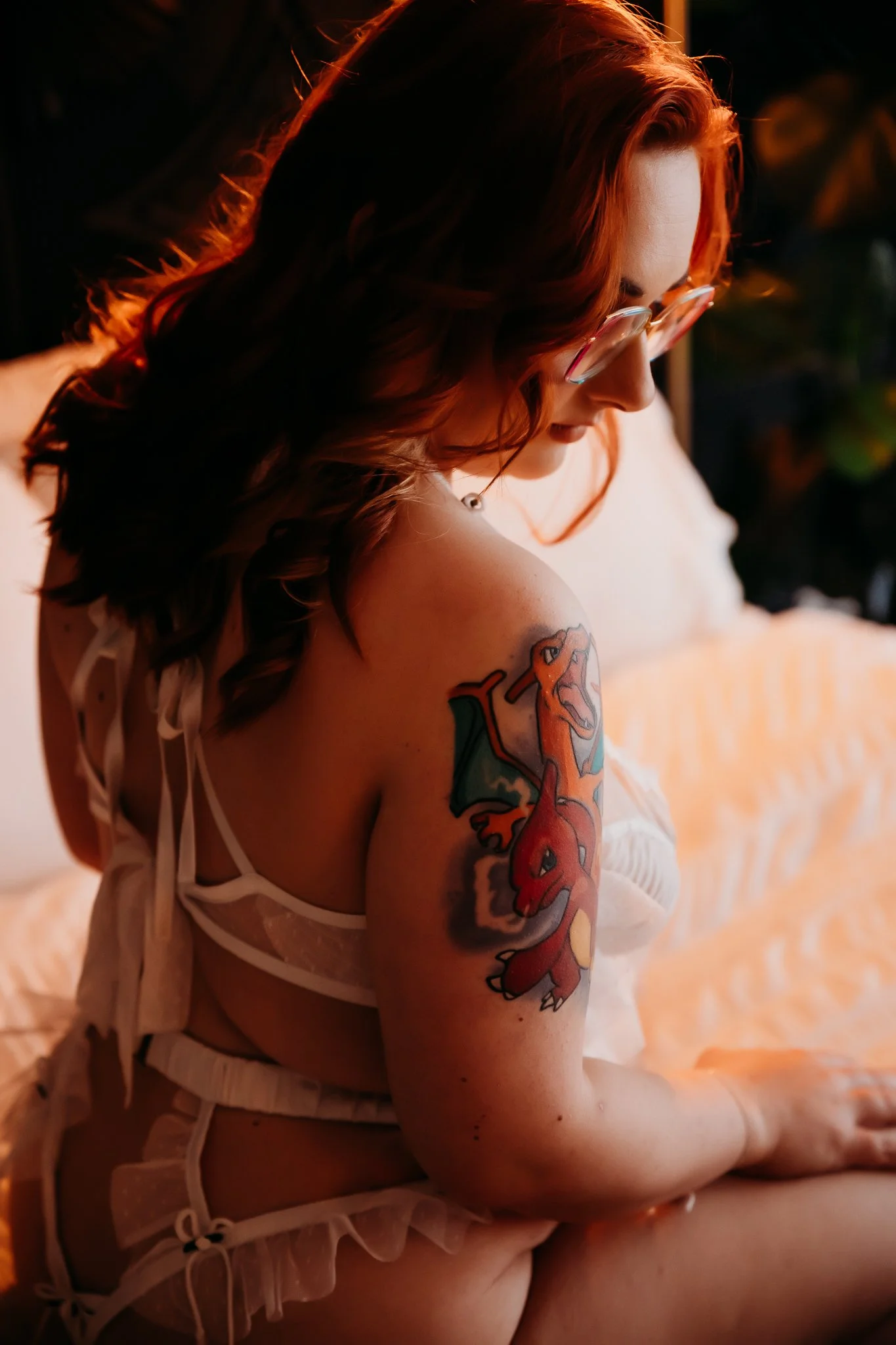 A woman with red hair and glasses sitting on a bed, looking down, showing a colorful dragon tattoo on her upper arm.