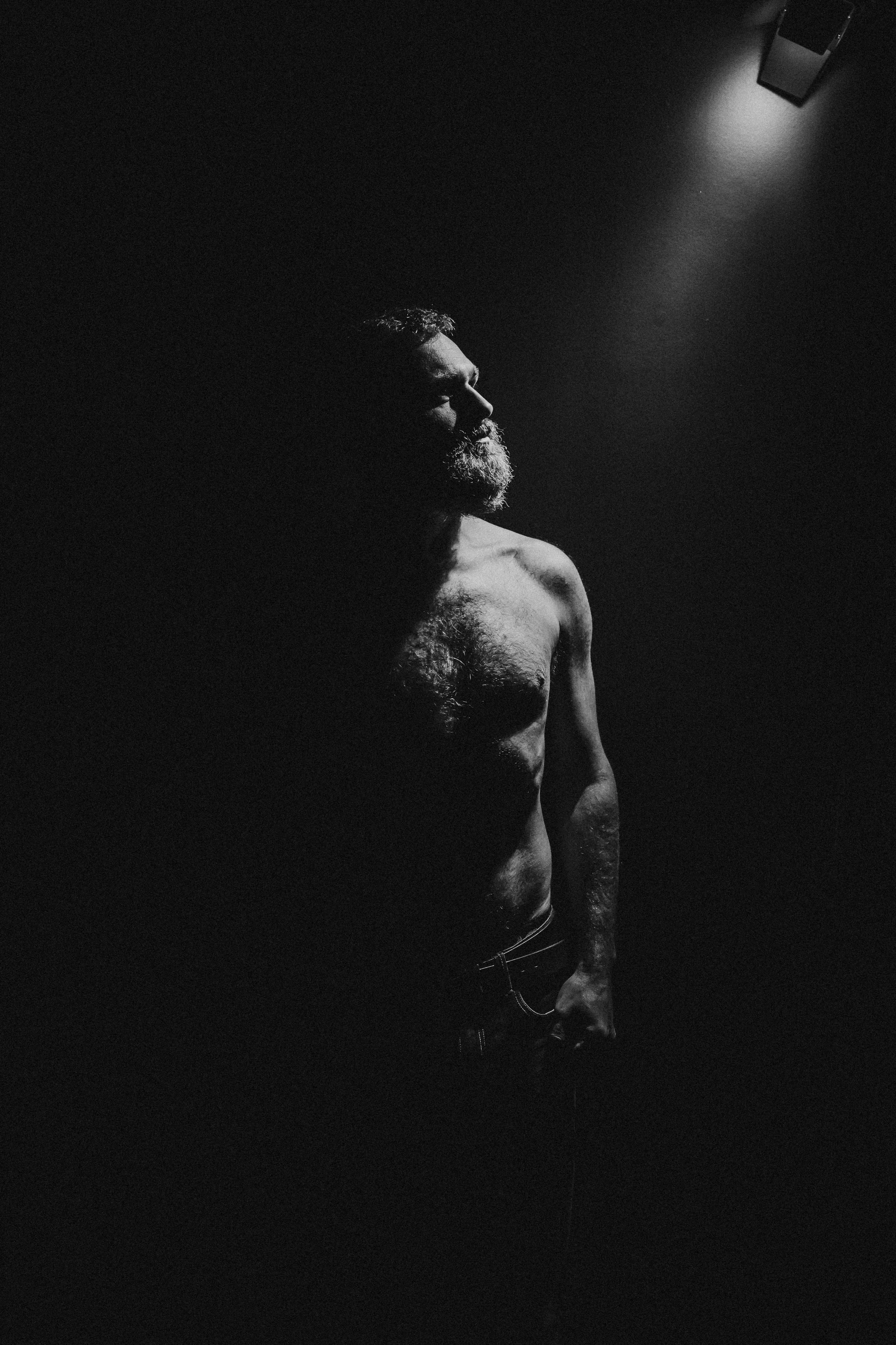 Black and white photo of a shirtless man with a beard standing against a dark background, illuminated by a ceiling light.
